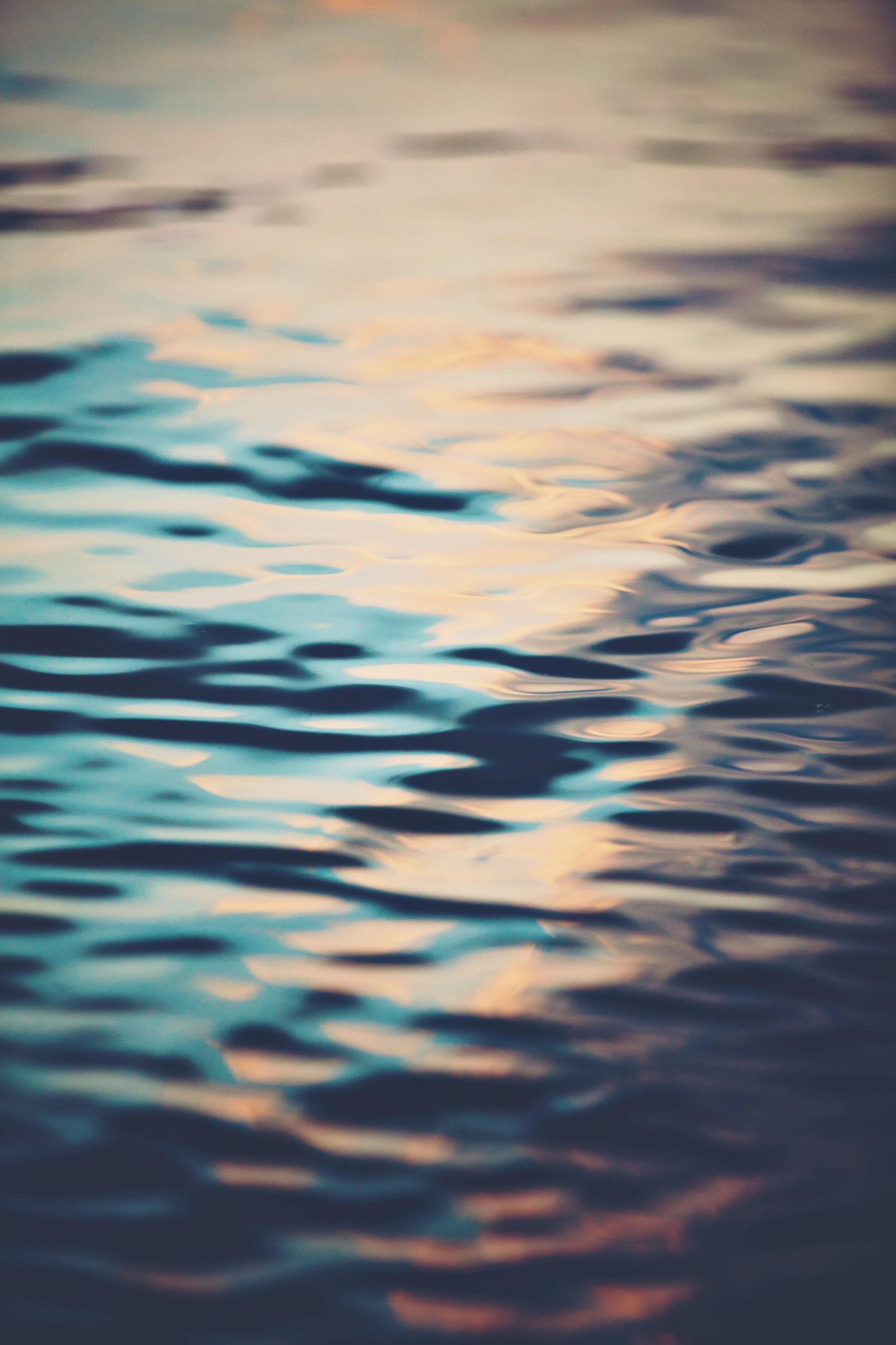 Softly blurred image of water with gentle ripples and pastel colors of the sky reflected on the surface.