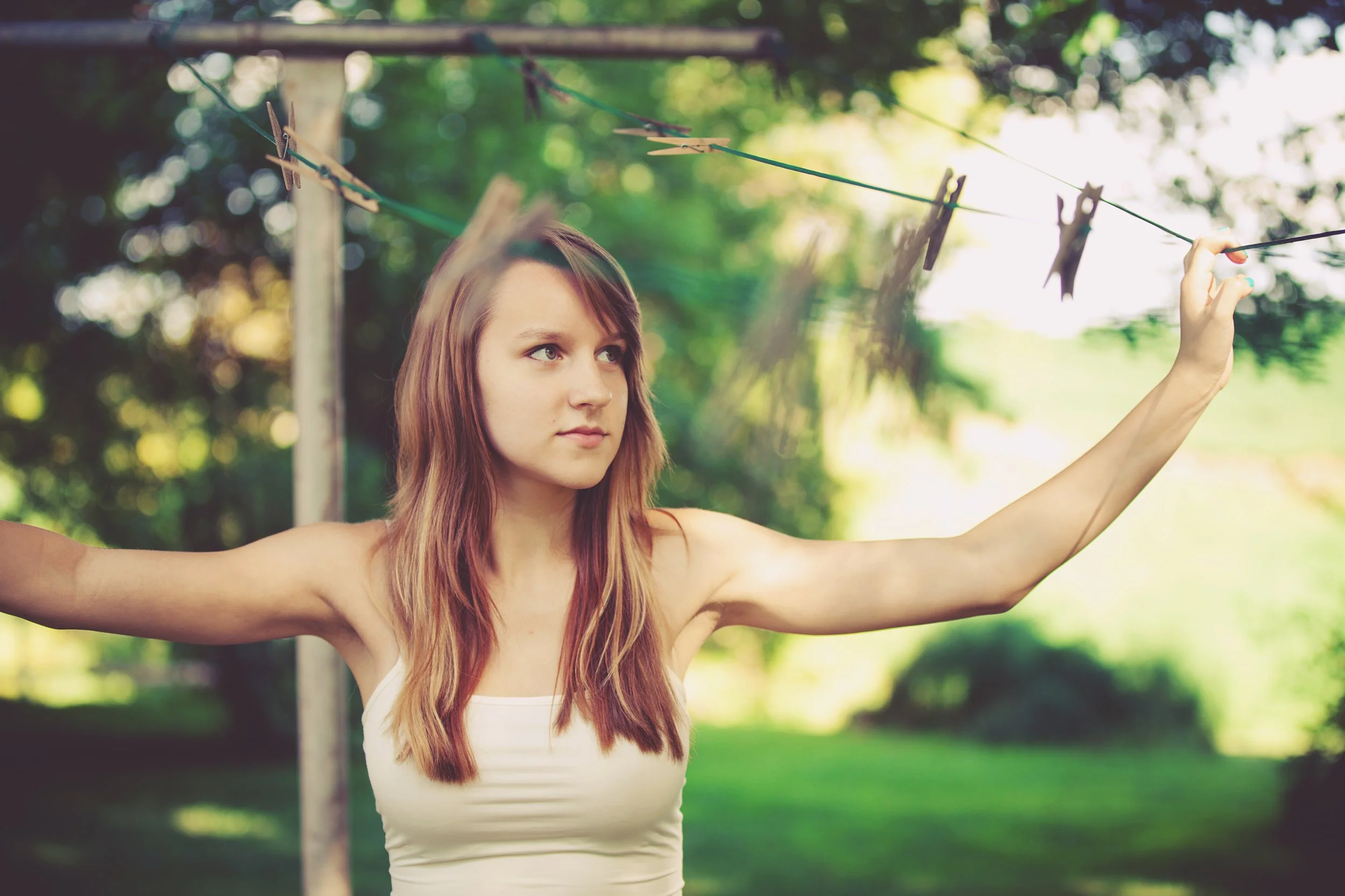 A young woman with long reddish-brown hair and fair skin hanging up laundry with clothespins on a clothesline outdoors in a green, leafy area.