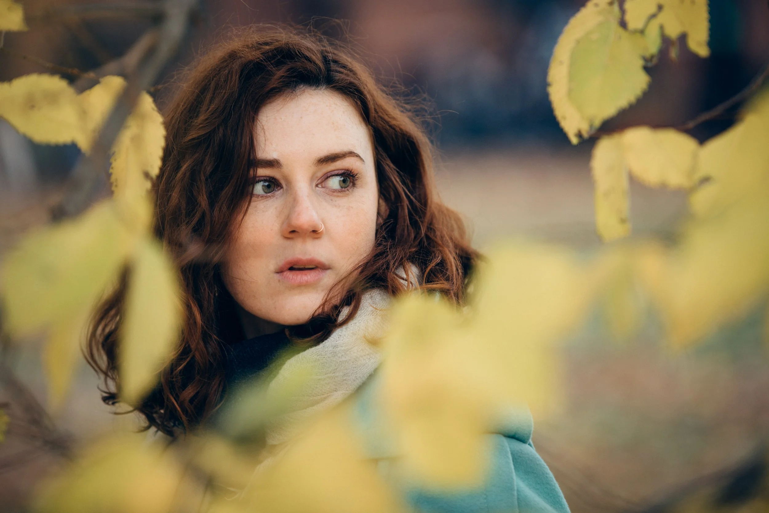 A young woman with curly brown hair and green eyes looking to her right, surrounded by yellow-green leaves outdoors.
