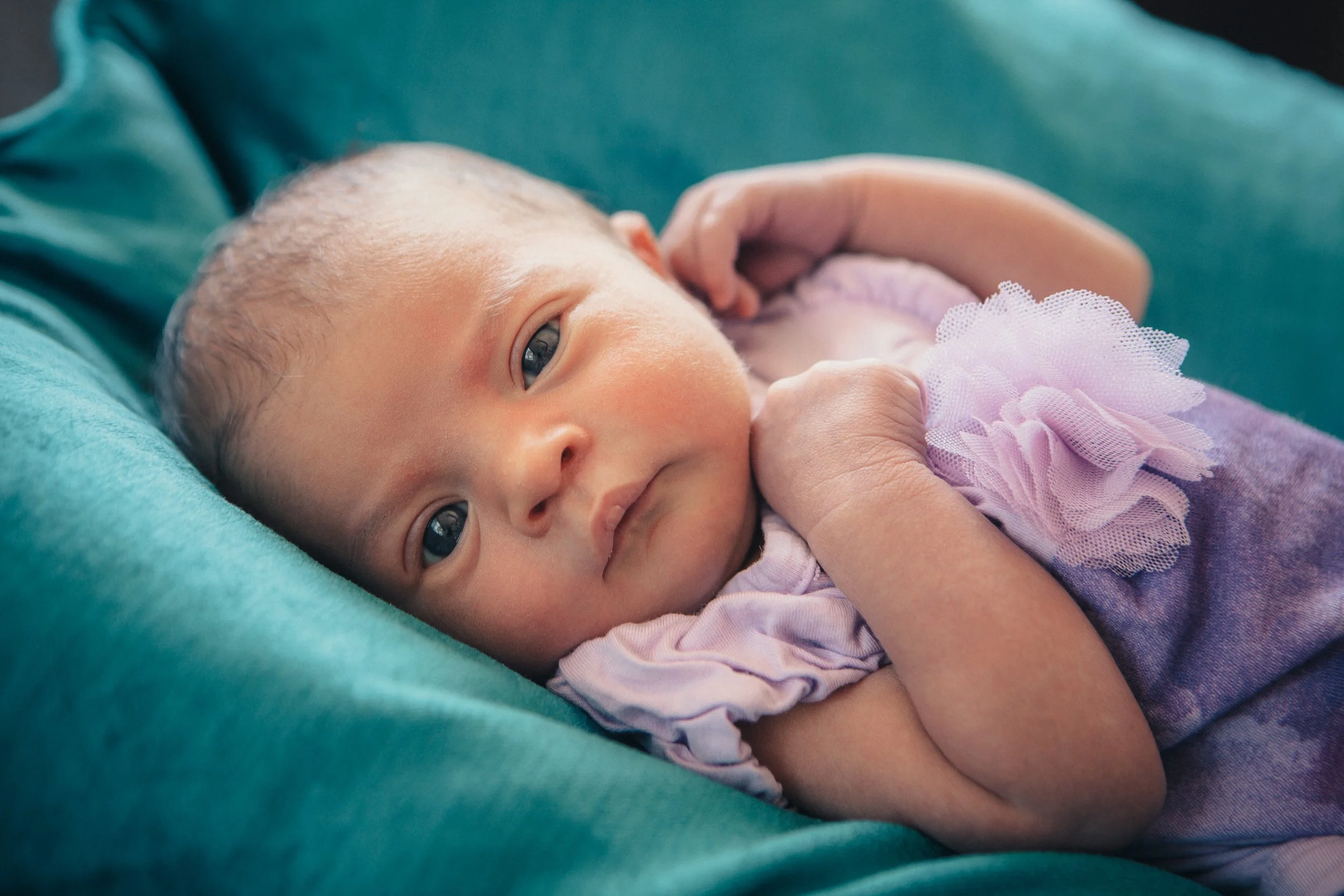 Close-up of a newborn baby lying on a person's arm, with the baby's gaze directed at the camera. The baby has a light complexion, light-colored eyes, and is wearing a pink outfit with a tulle accent. The background includes teal and purple fabric.