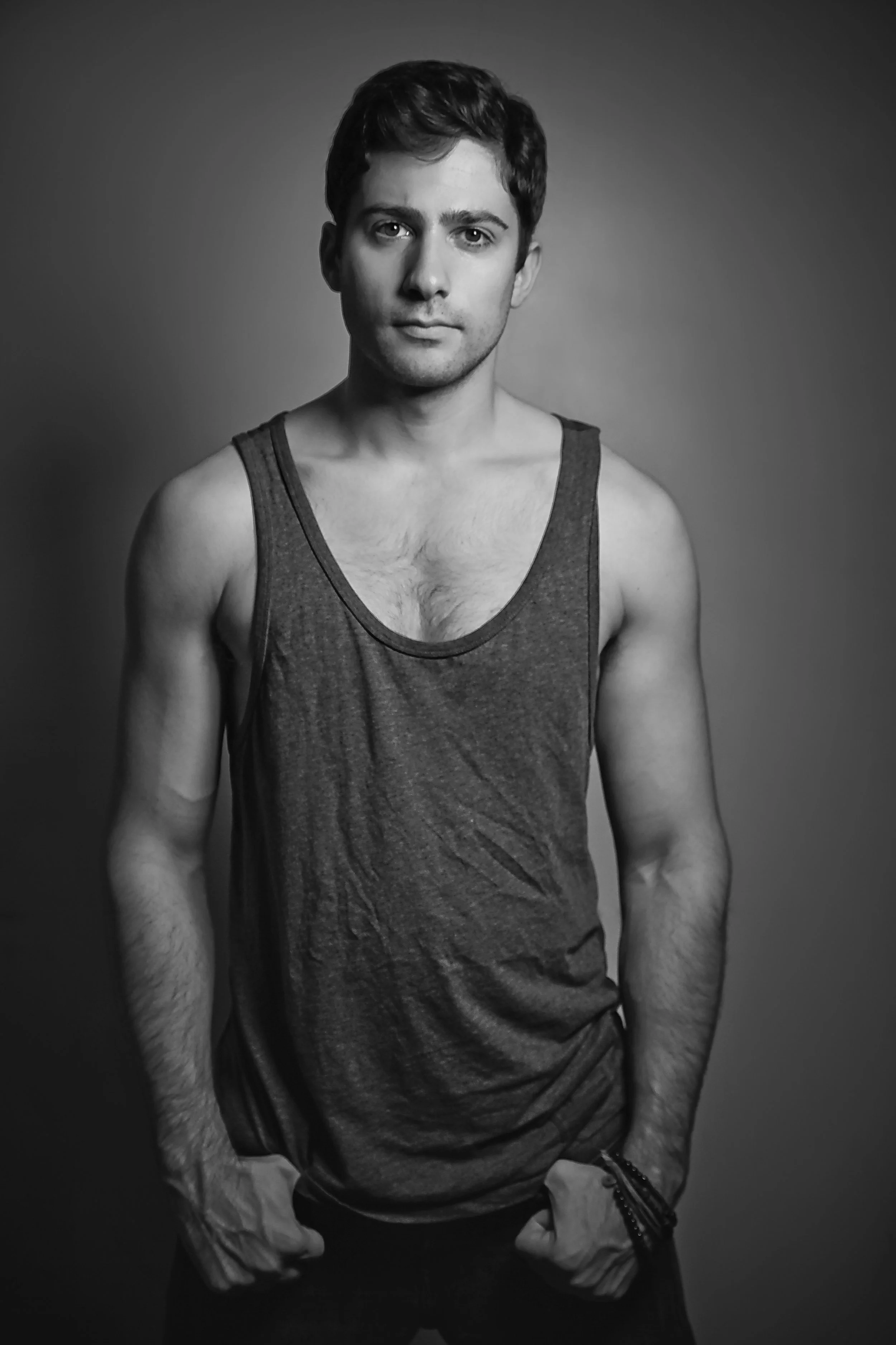 Black and white photograph of a young man with short dark hair, wearing a sleeveless tank top, standing against a plain background. He has a serious expression and his hands are resting in his pockets, with a bracelet visible on his left wrist.