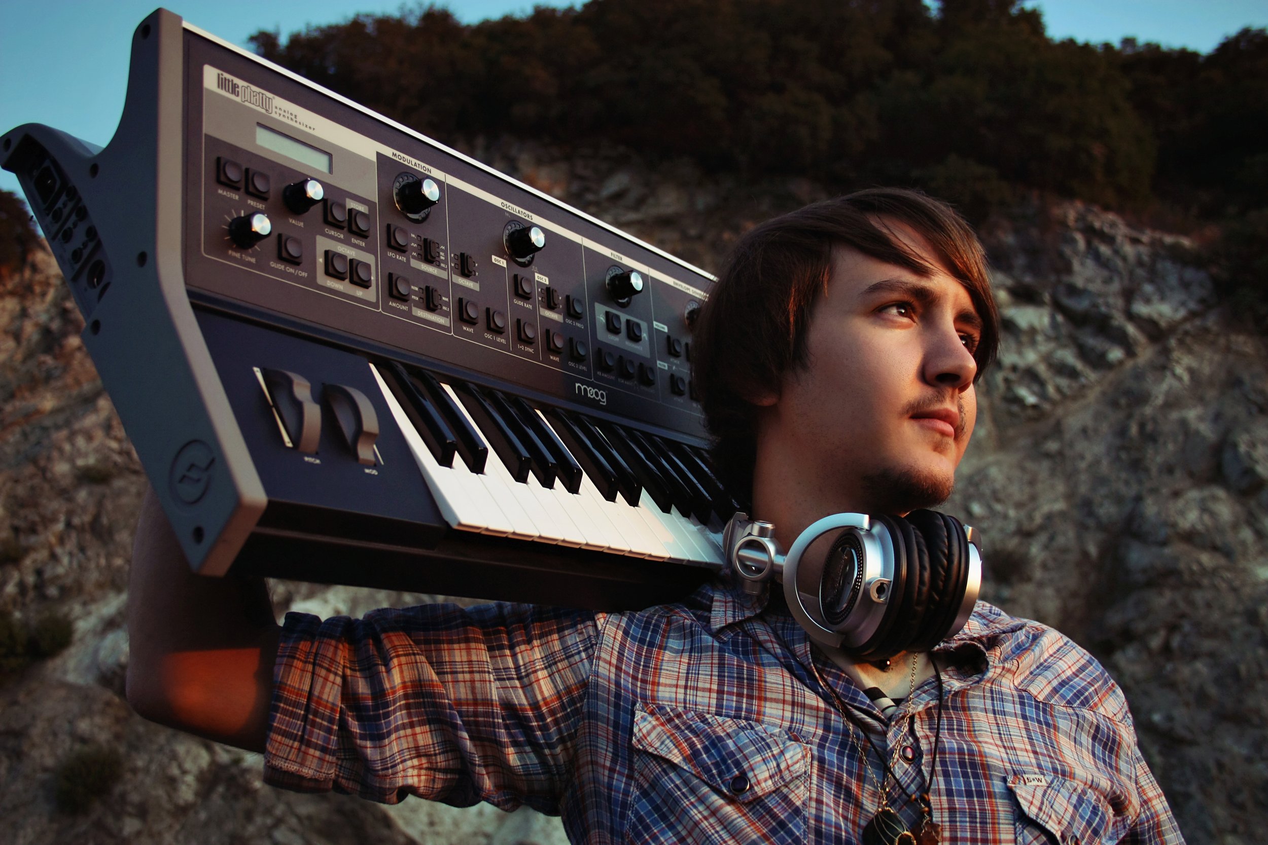 A young man outdoors with a mountain landscape in the background, carrying a keyboard on his shoulder and wearing headphones around his neck.
