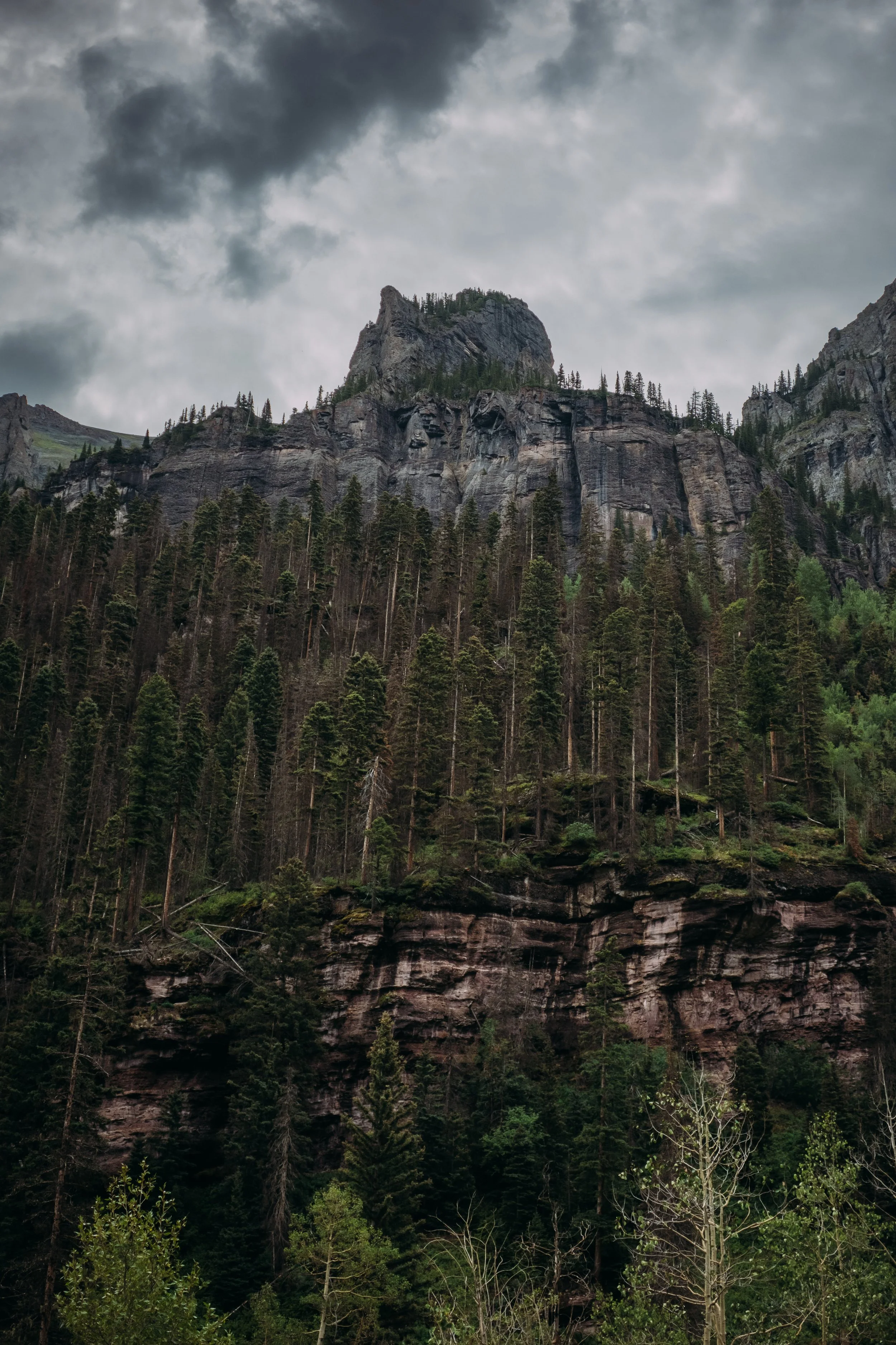 A mountain with a steepling peak, surrounded by dense green pine forest, under a cloudy sky.