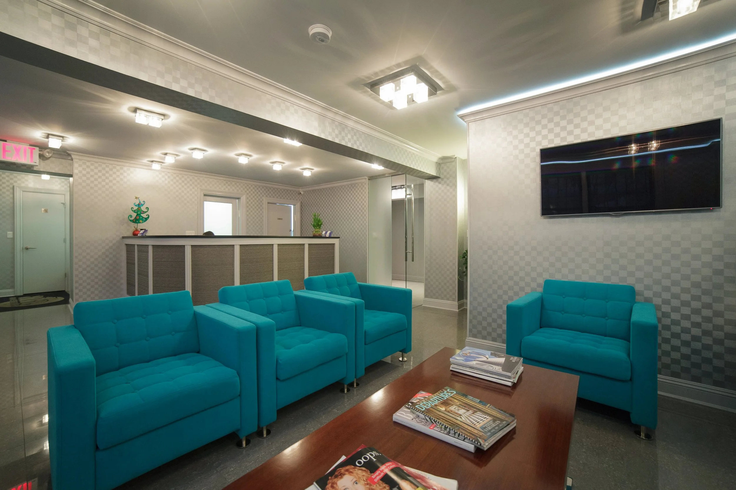 A waiting area with turquoise cushioned chairs, a wooden coffee table with magazines, and a wall-mounted flat-screen TV. The background shows a reception desk with holiday decorations, and the walls are covered in light-colored checkered wallpaper wi
