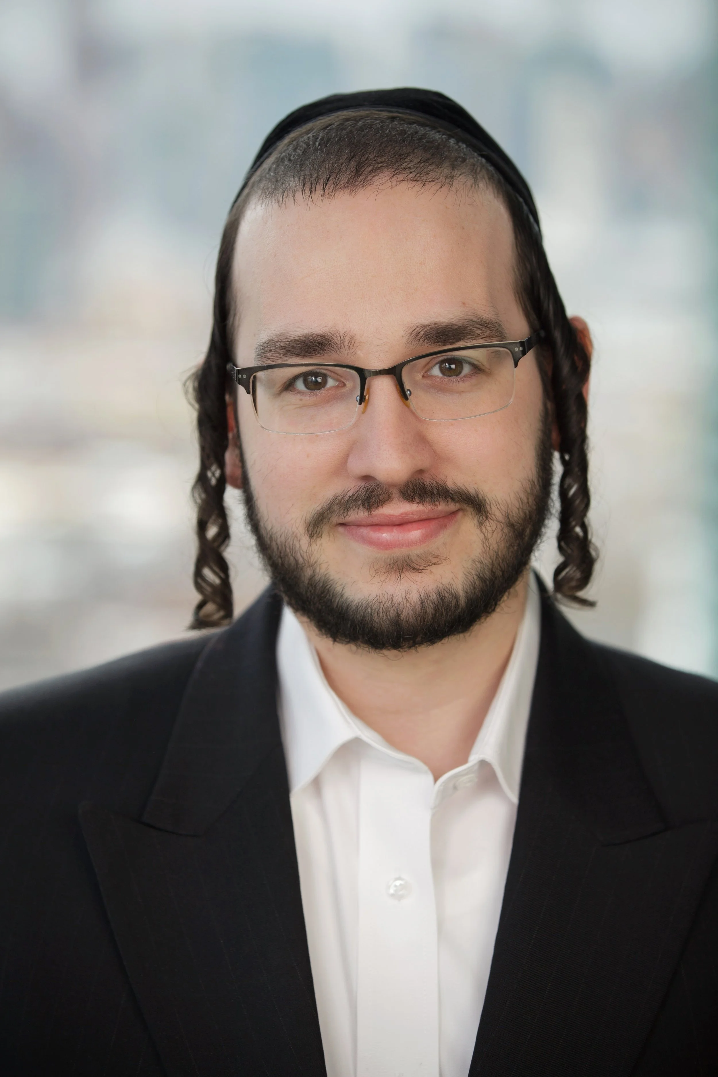 A man dressed in a black suit, white shirt, and glasses with curly hair and a beard, smiling for the photo.