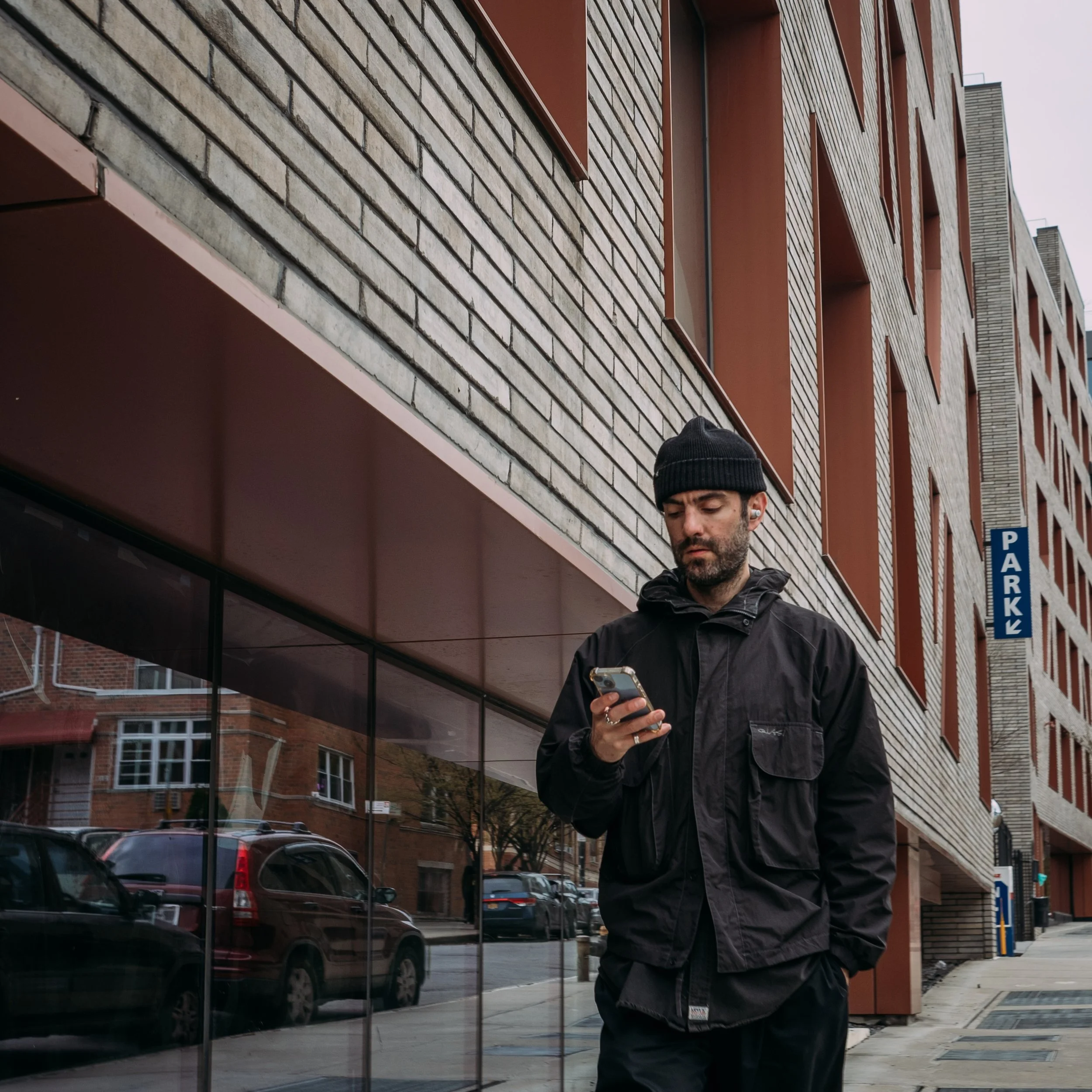 A man in a black jacket and beanie hat standing outdoors in front of a building, looking at his smartphone, with parked cars, trees, brick buildings, and a sign reading 'Parkw' nearby.