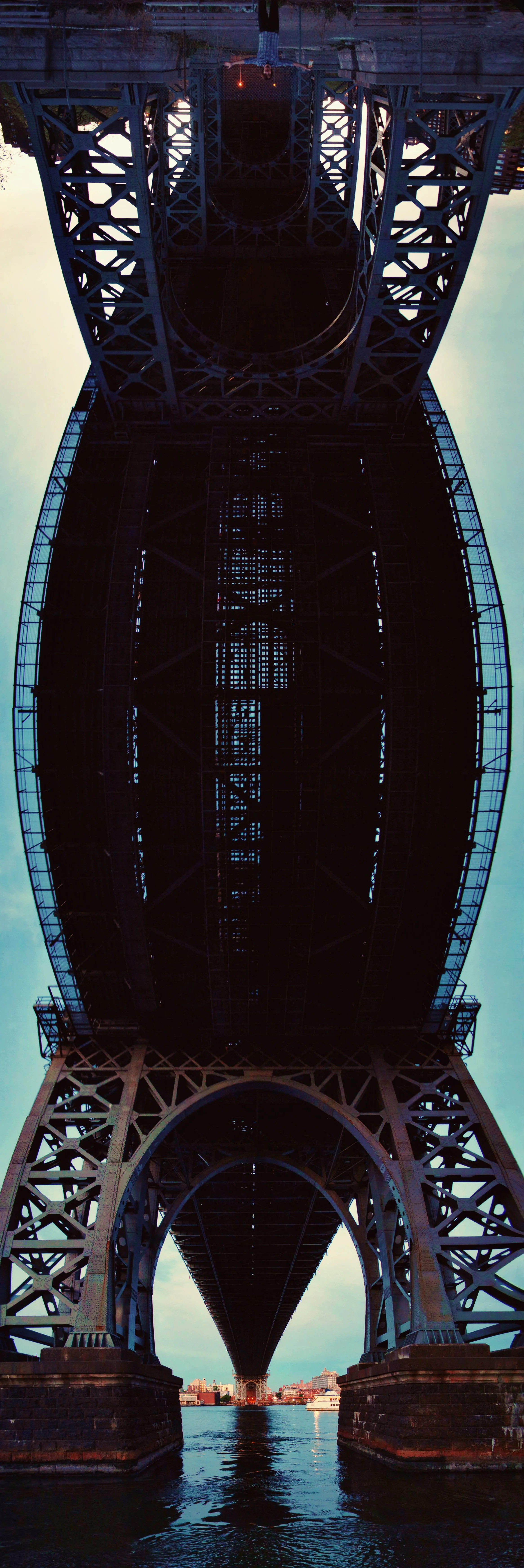 Underneath view of a large suspension bridge over a body of water, showing its intricate steel framework and girders, with a city skyline in the background.