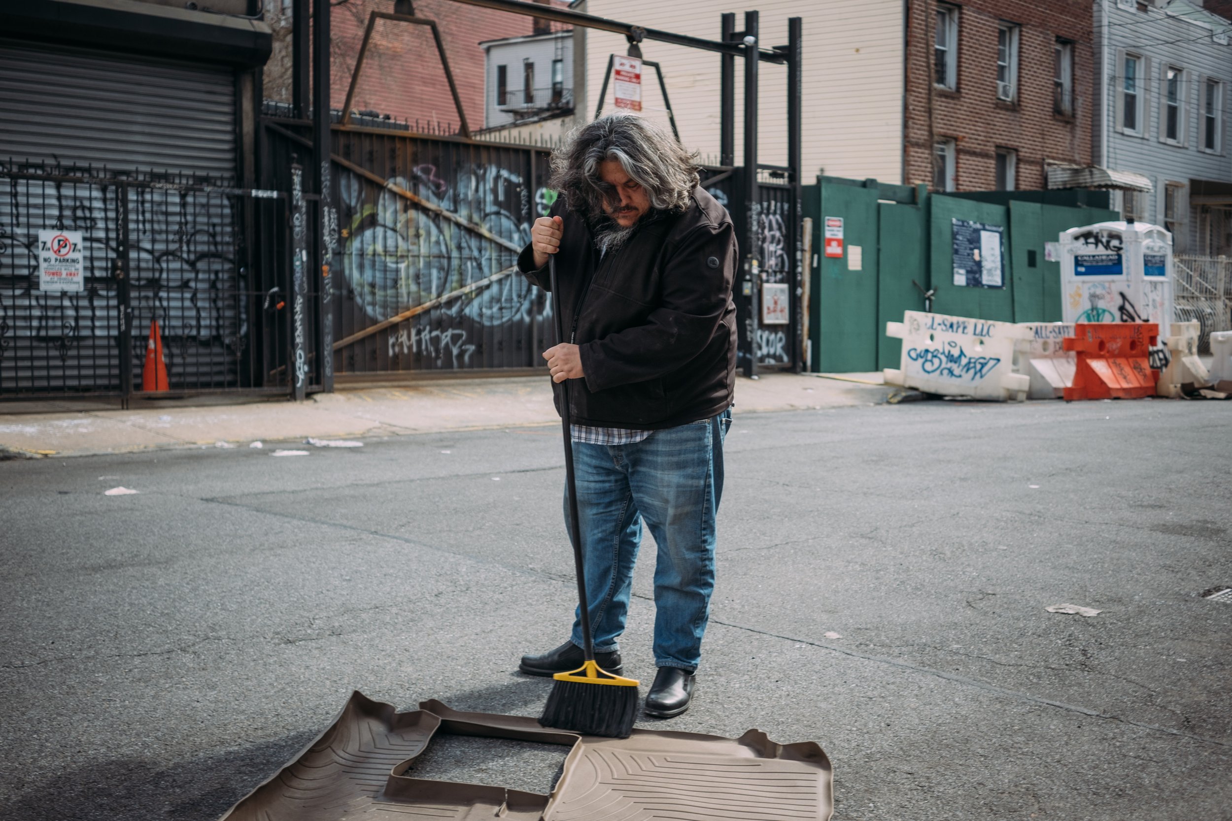 A man with gray, curly hair and a beard is sweeping the street with a broom while standing on an urban sidewalk. He is wearing a black jacket, plaid shirt, jeans, and black boots. There are graffiti-covered gates and graffiti-covered walls with buildings in the background, along with construction barriers and signs.