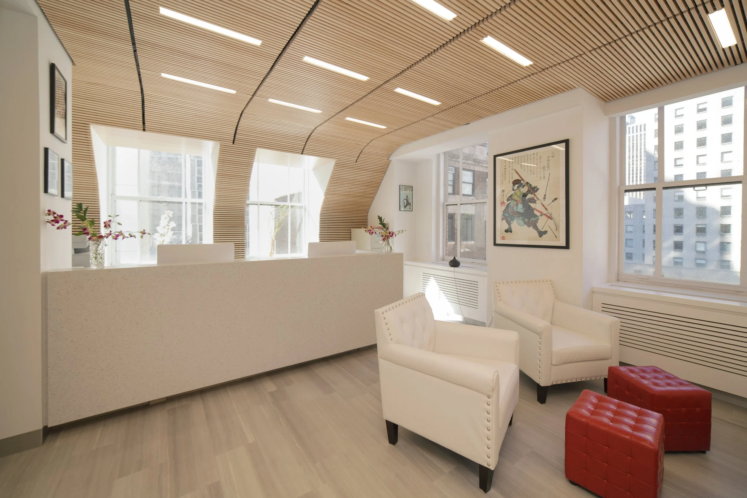 Modern waiting area with white chairs, red ottomans, and framed artwork on the wall, illuminated by natural light through large windows.