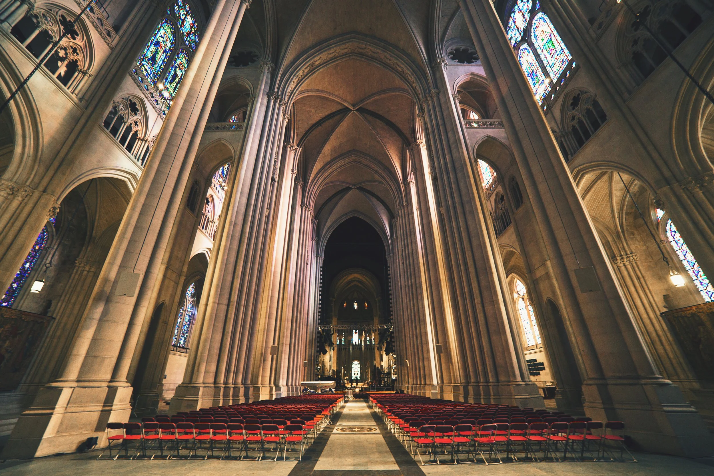 Interior of a Gothic-style cathedral with high vaulted ceilings, stained glass windows, and rows of red chairs.