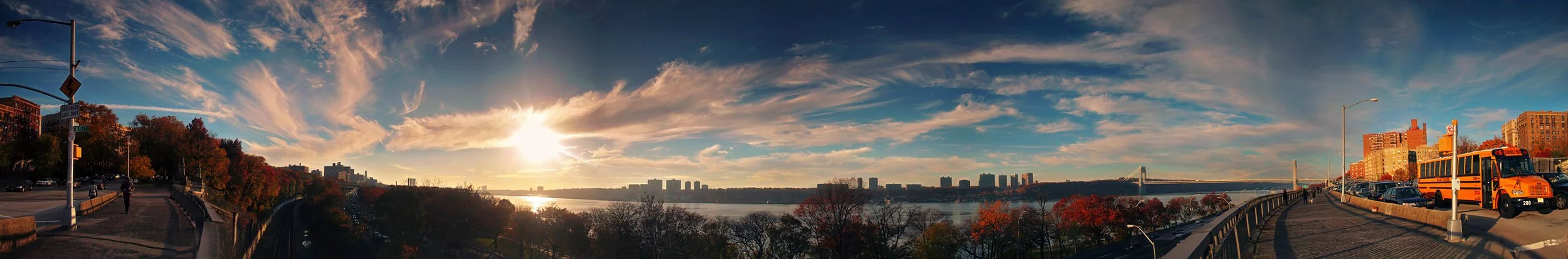 Panoramic view of a city skyline during sunset with a river in the middle, a bridge in the distance, trees with fall foliage, a walkway with a person walking, parked cars, and a bus on the right side.