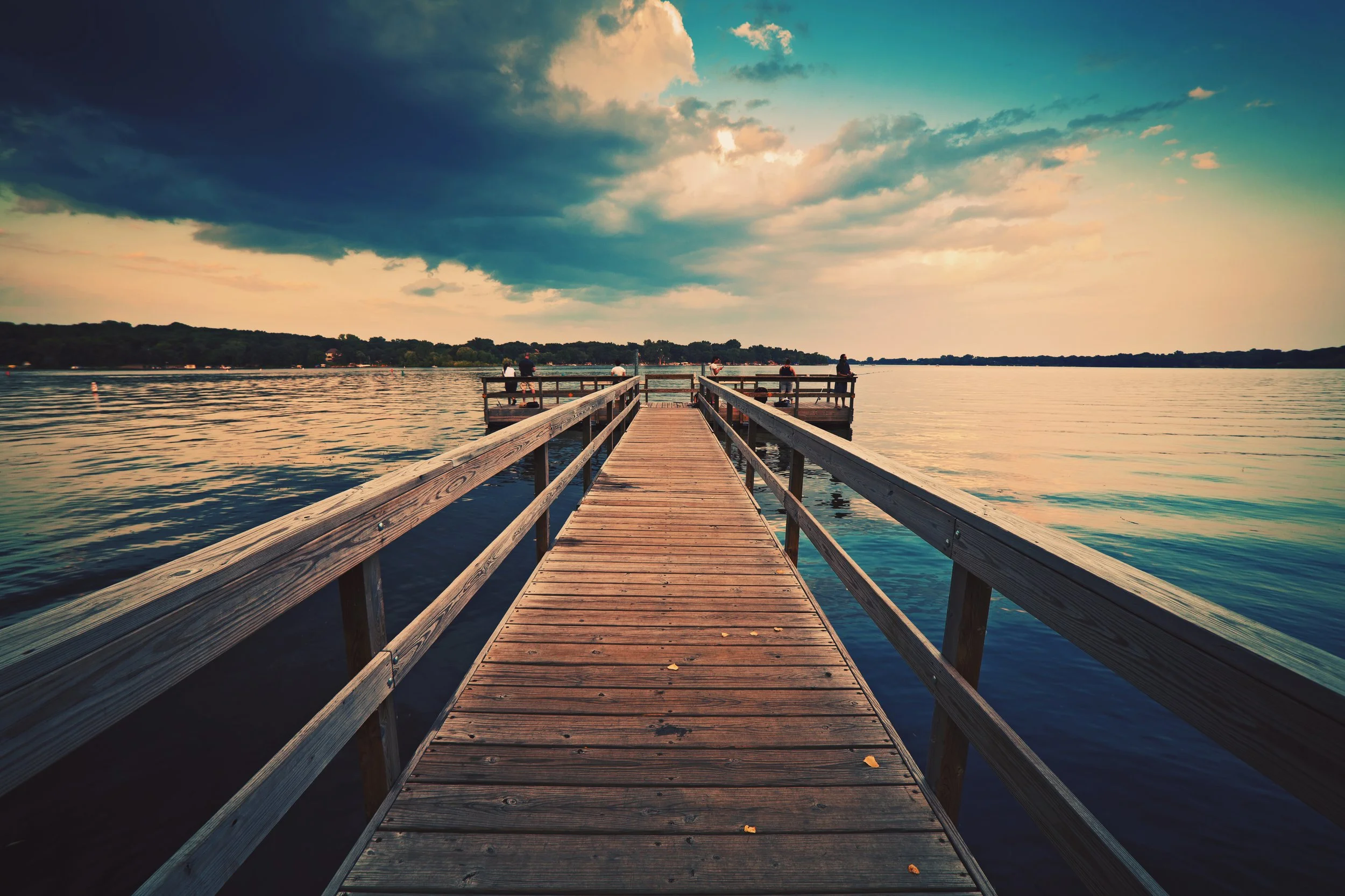 A wooden pier extends into a calm body of water under a dramatic sky with dark and light clouds during sunset or sunrise, with a few people fishing along the pier.