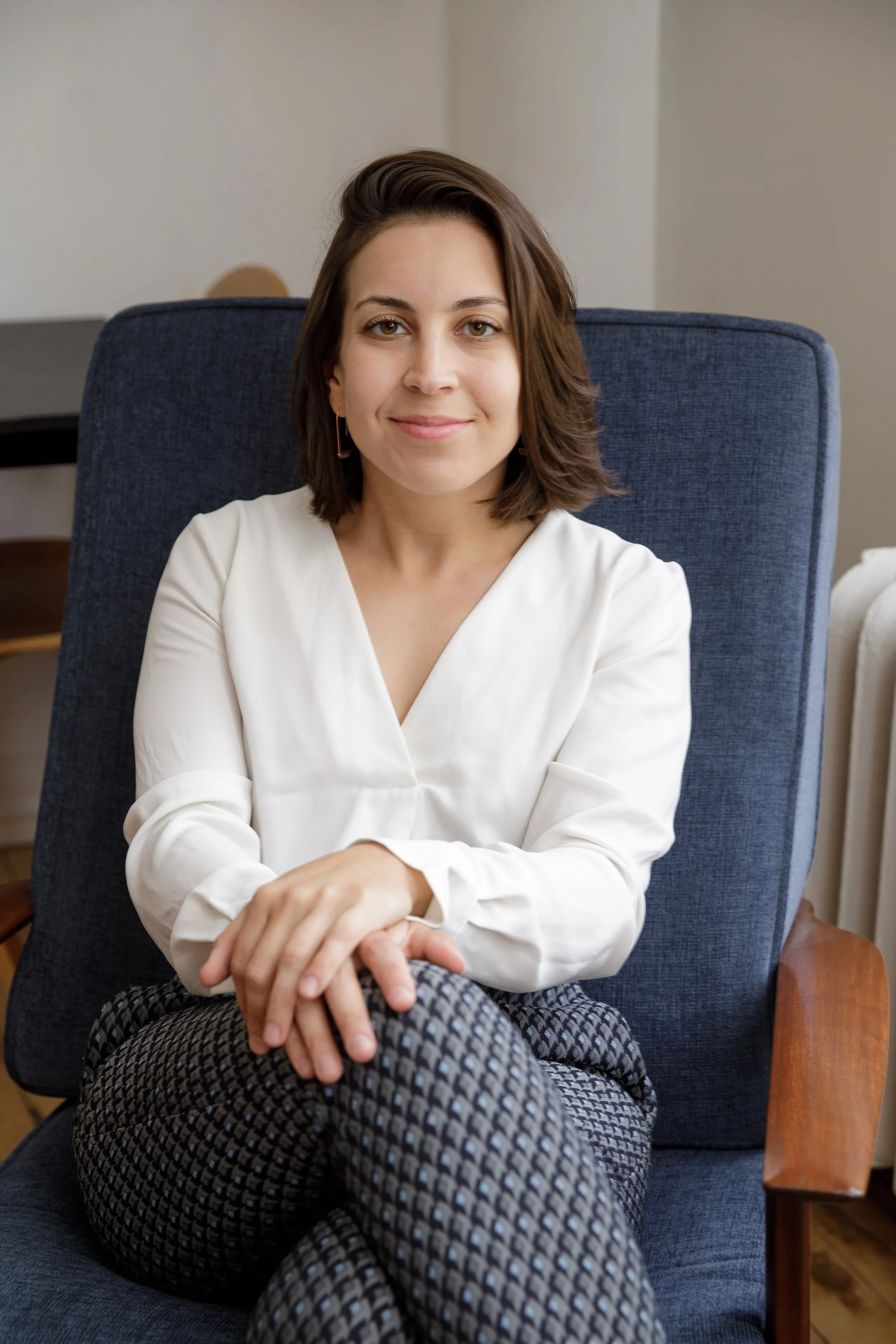 A woman with shoulder-length brown hair sitting on a blue upholstered armchair, smiling softly, wearing a white blouse and patterned pants, with a wooden side table visible in the background.