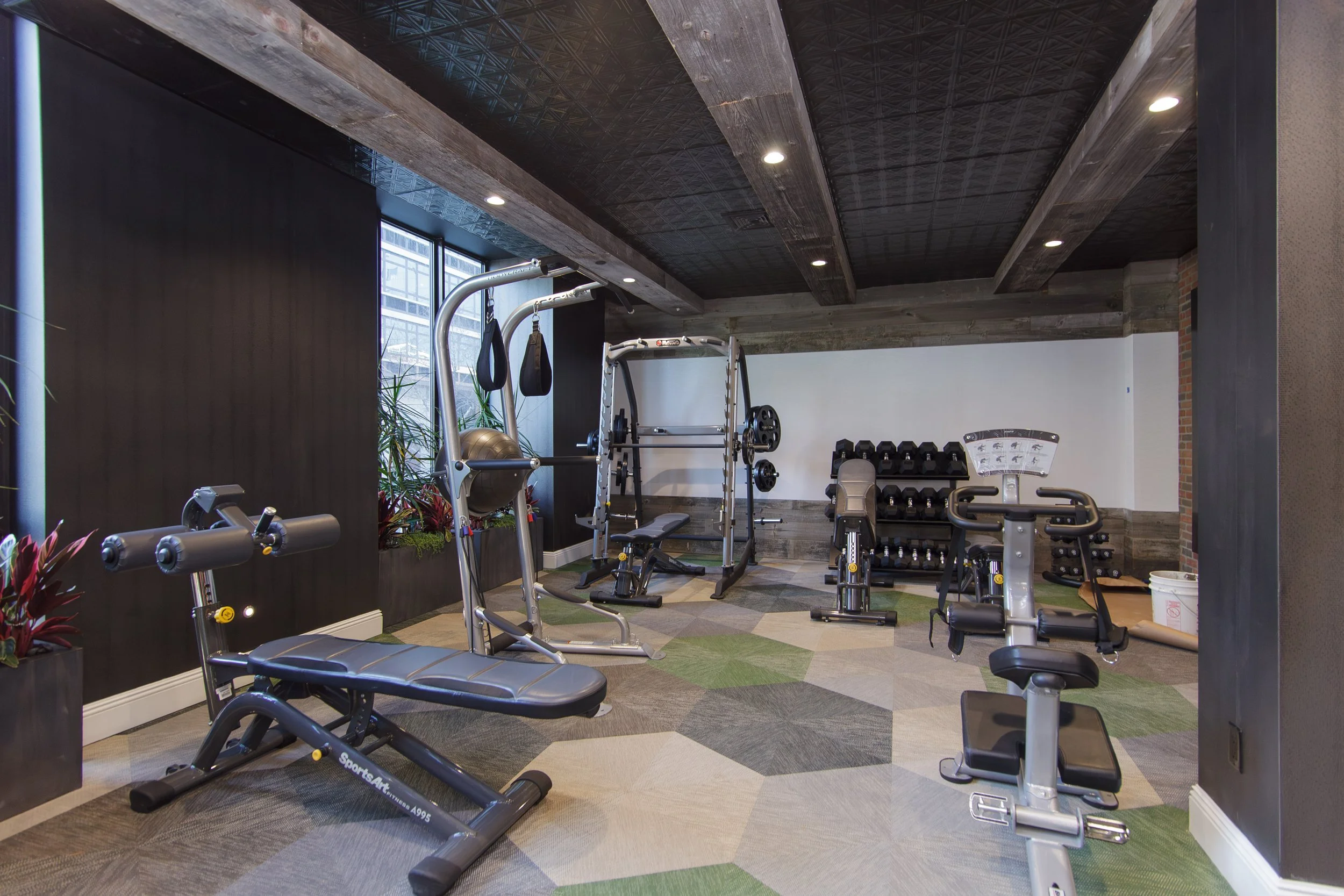 A modern gym with various exercise equipment including a treadmill, weight bench, dumbbells, and resistance machines. The gym has stone and brick accents, large windows, and a patterned carpeted floor.