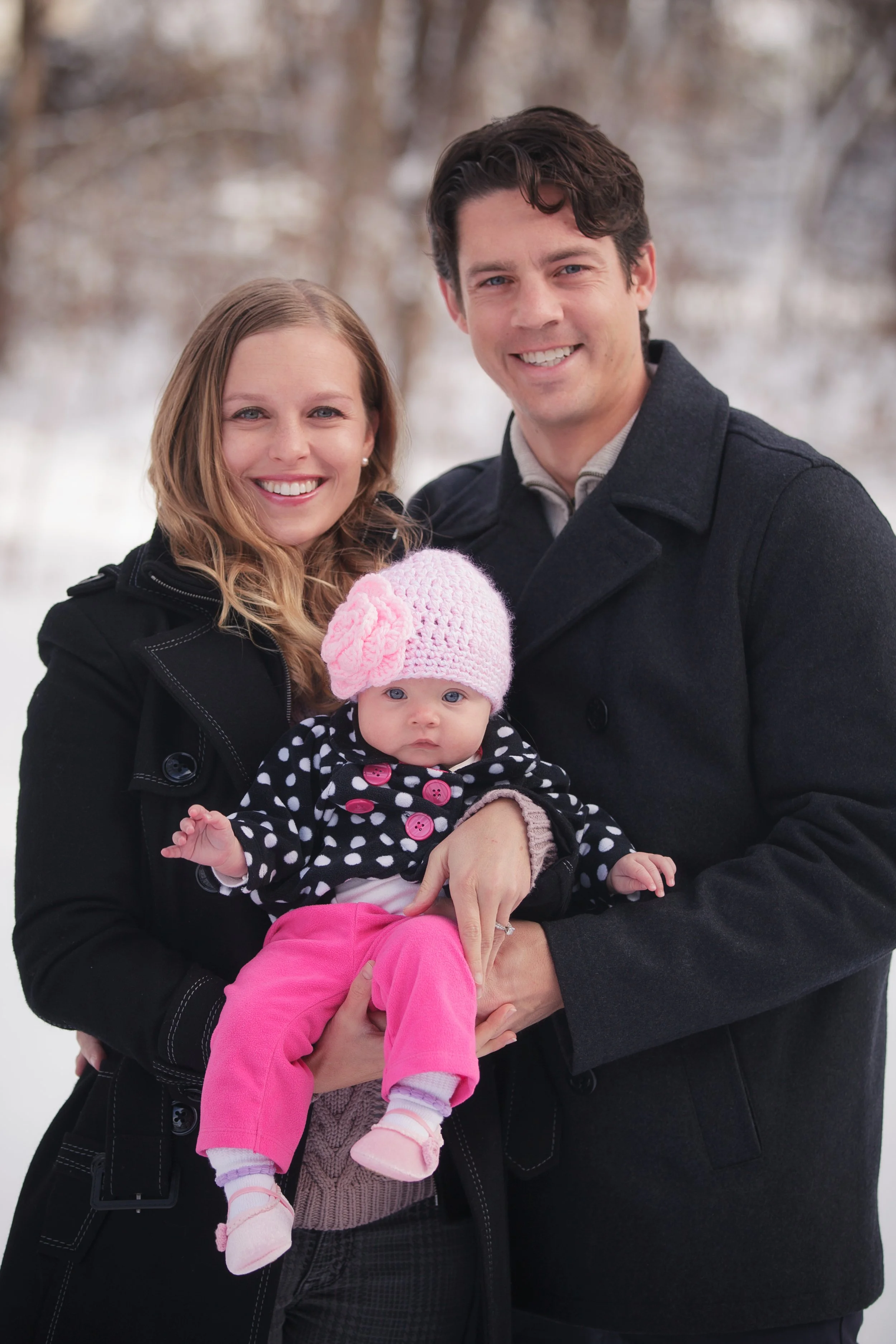 A family of three, including a woman, a man, and a baby girl, standing outdoors in winter, smiling at the camera with a snowy background.