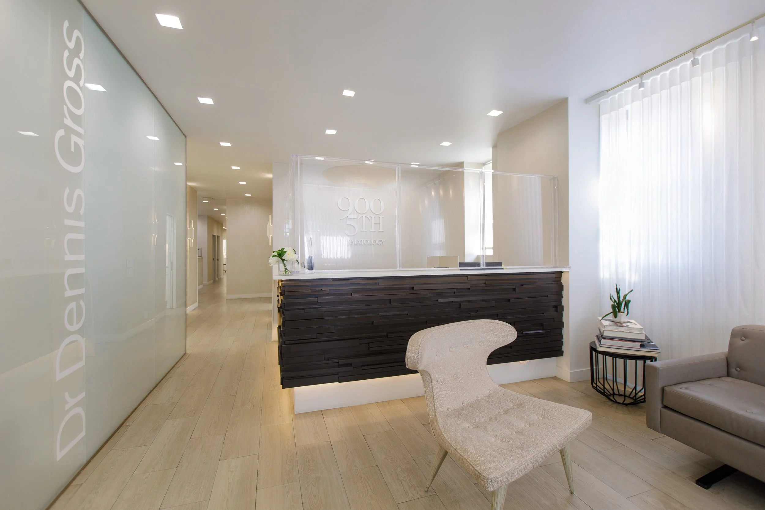 Clinic reception area with a modern black and white front desk, white walls, a plant on the right side, a stack of magazines beside the plant, two chairs, and built-in ceiling lights.