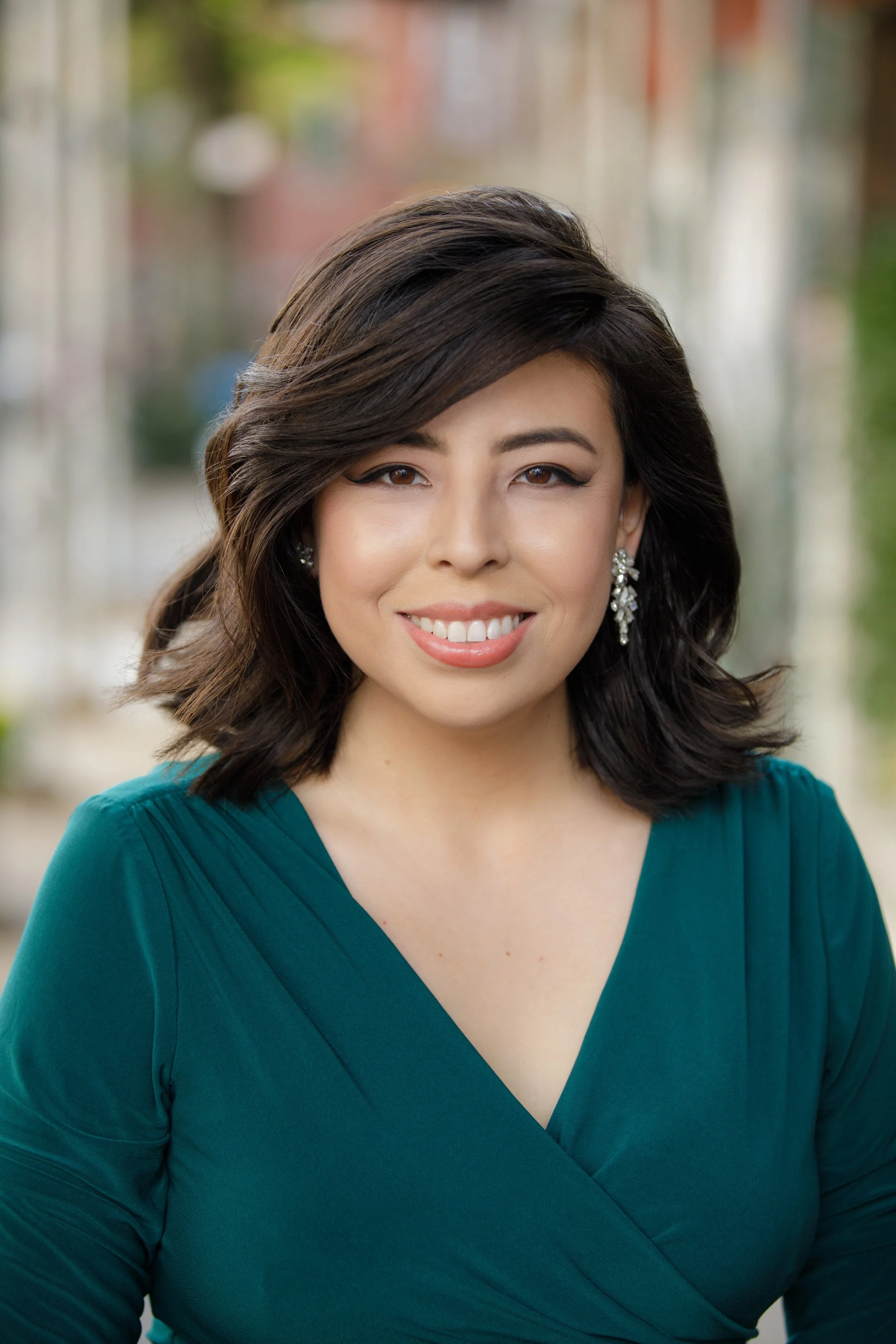 Close-up of a smiling woman with dark brown hair styled in soft waves, wearing diamond earrings and a teal V-neck dress, standing outdoors with a blurred city street in the background.