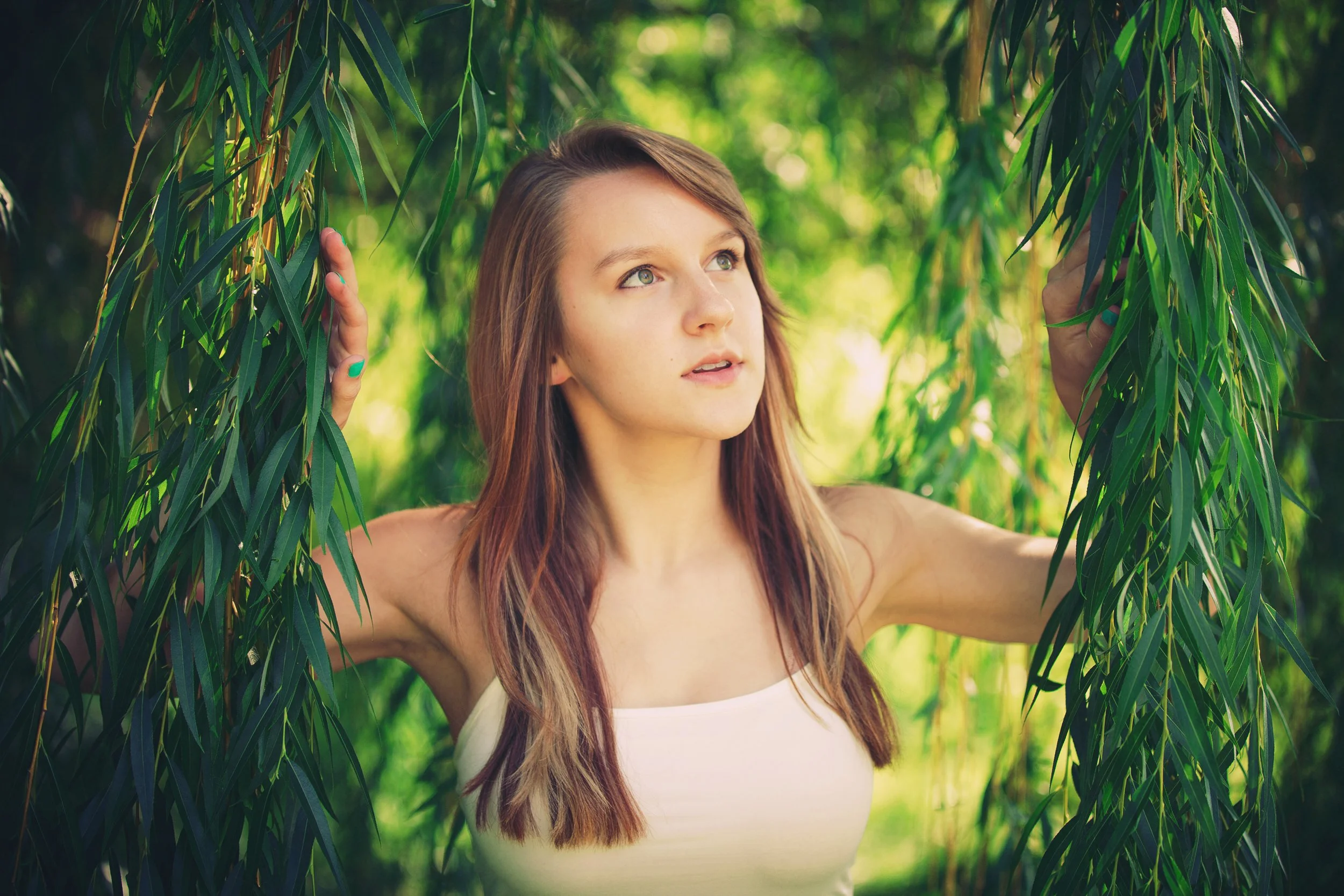 A young woman with long brown hair and green eyes standing among green leafy trees, touching the branches with her hands, wearing a white strapless top.