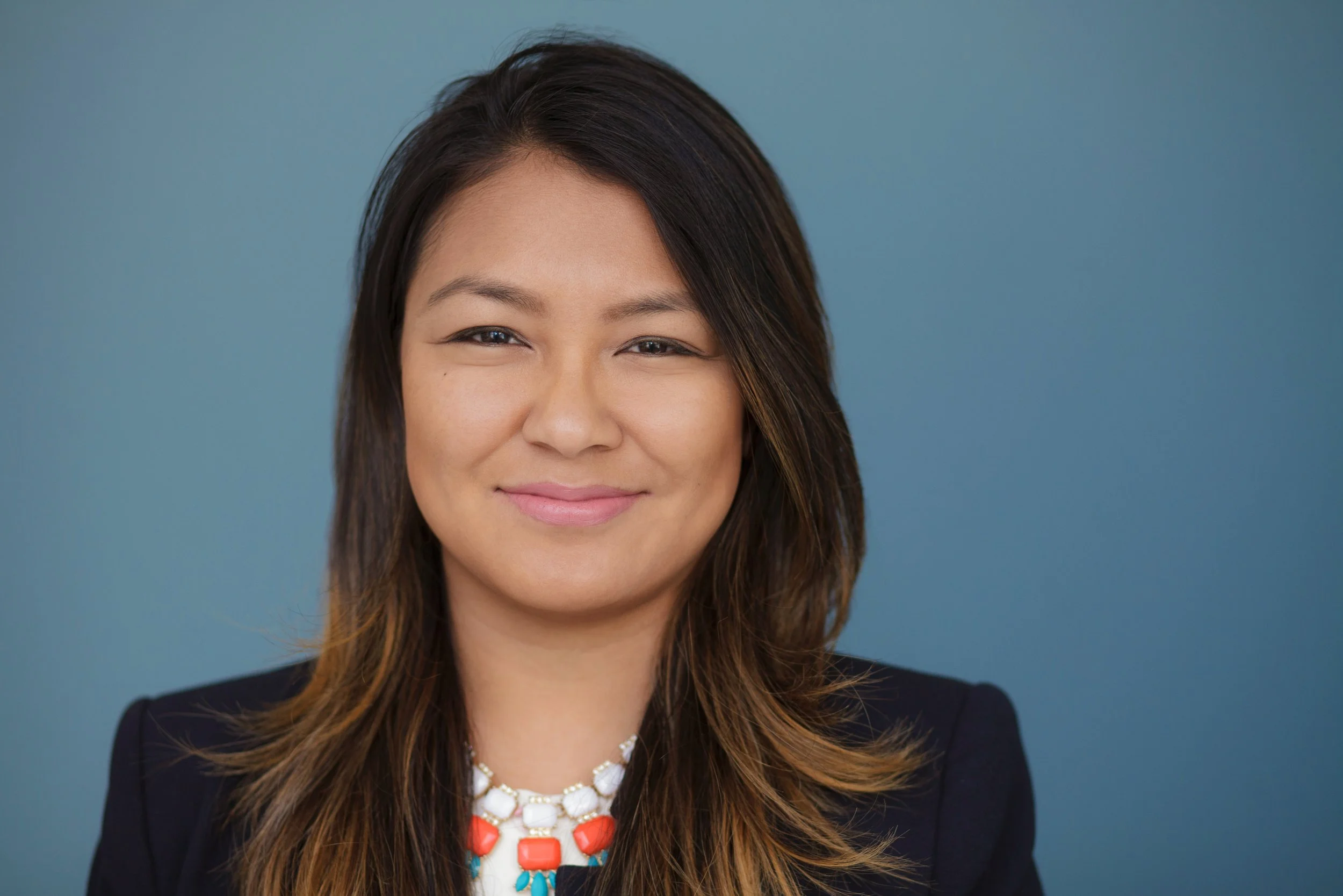 Close-up of a smiling woman with long brown hair wearing a black blazer and aStatement necklace with large white, coral, and turquoise beads, against a solid blue background.