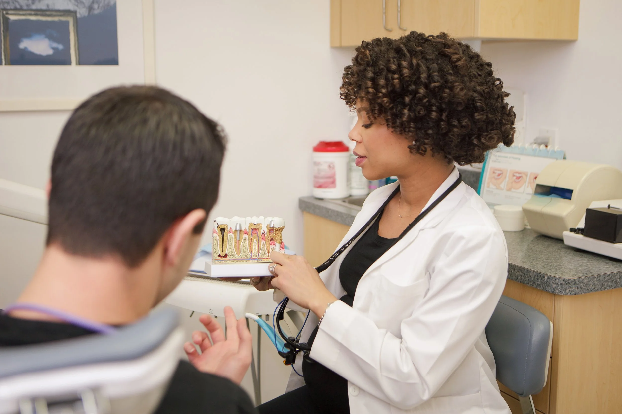 A dental professional wearing a white coat and stethoscope shows a model of teeth to a patient at a dental office.