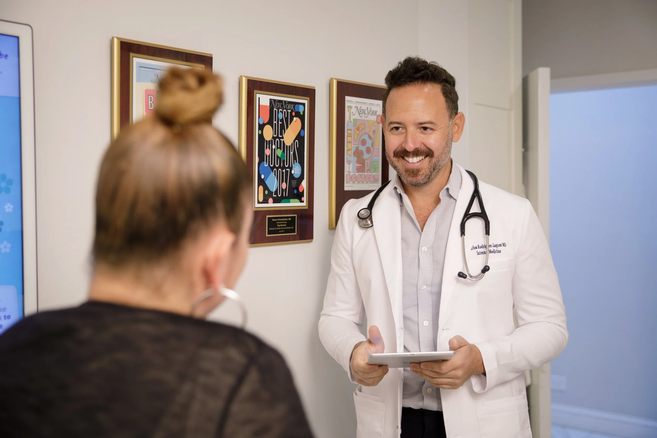 A male doctor with a stethoscope around his neck smiling and speaking to a patient in a medical office or clinic.