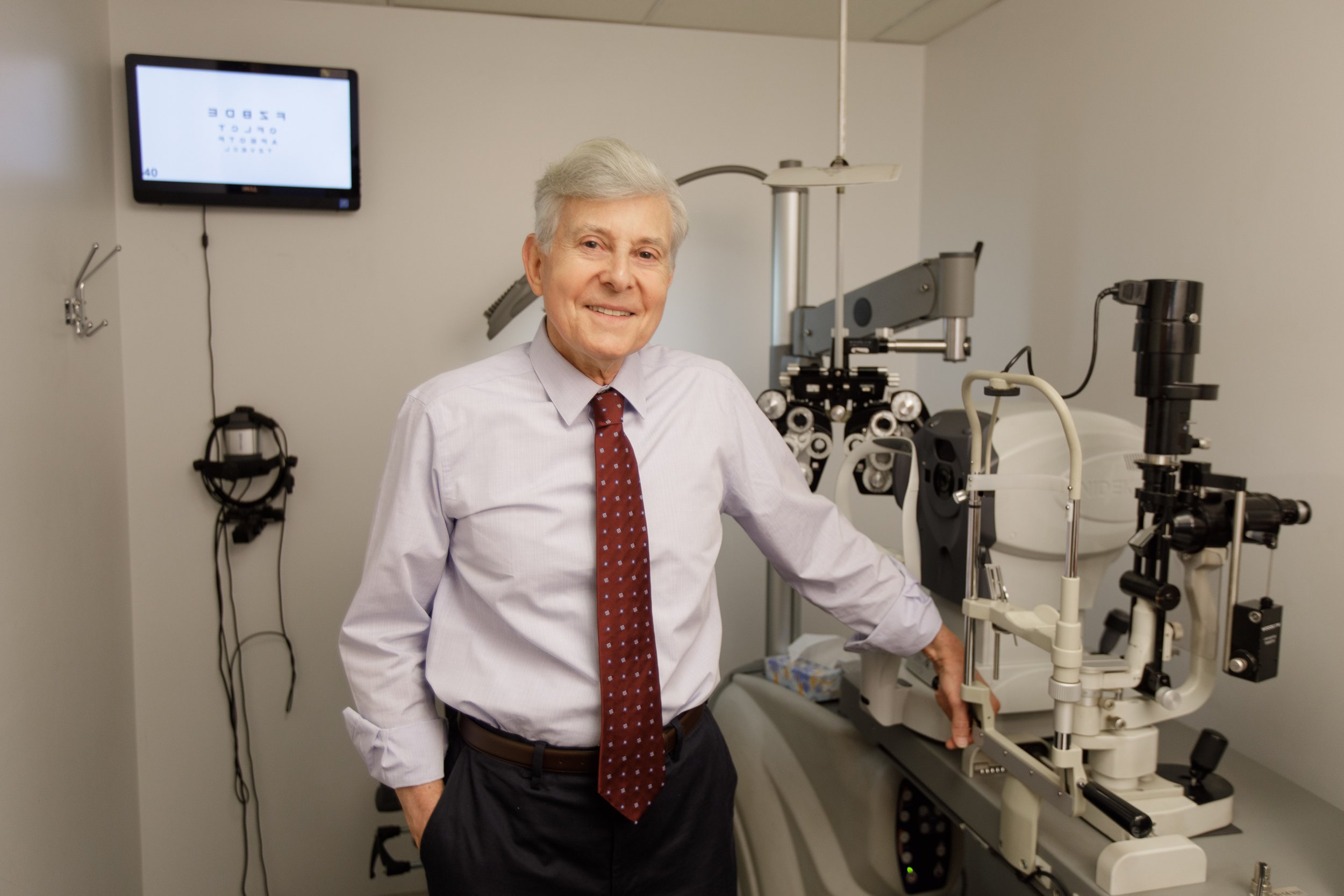A smiling elderly man in a white shirt and red tie standing next to ophthalmology equipment in a medical examination room.