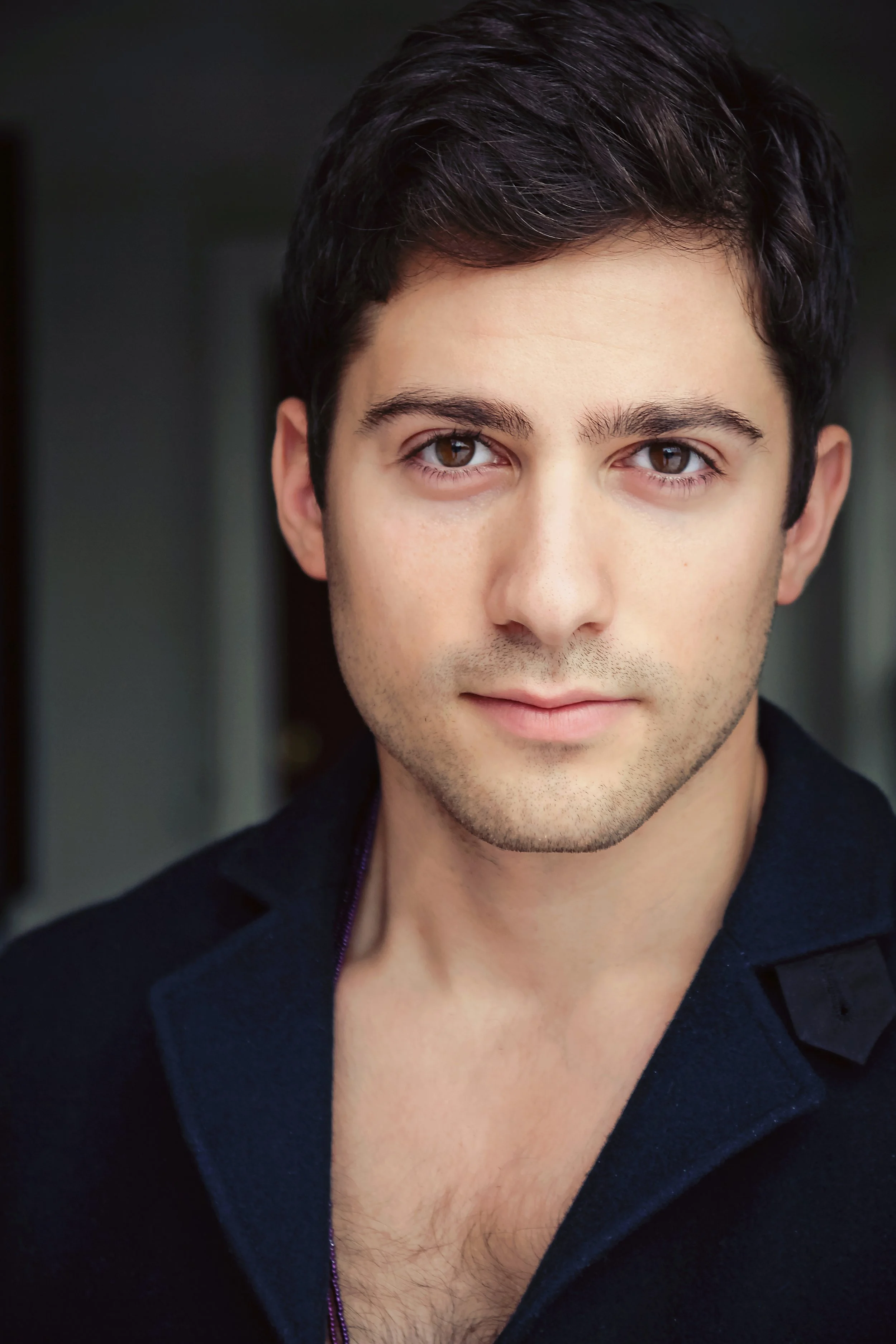 Close-up portrait of a young man with dark brown hair, brown eyes, fair skin, and slight facial hair, wearing a dark jacket, looking directly at the camera.