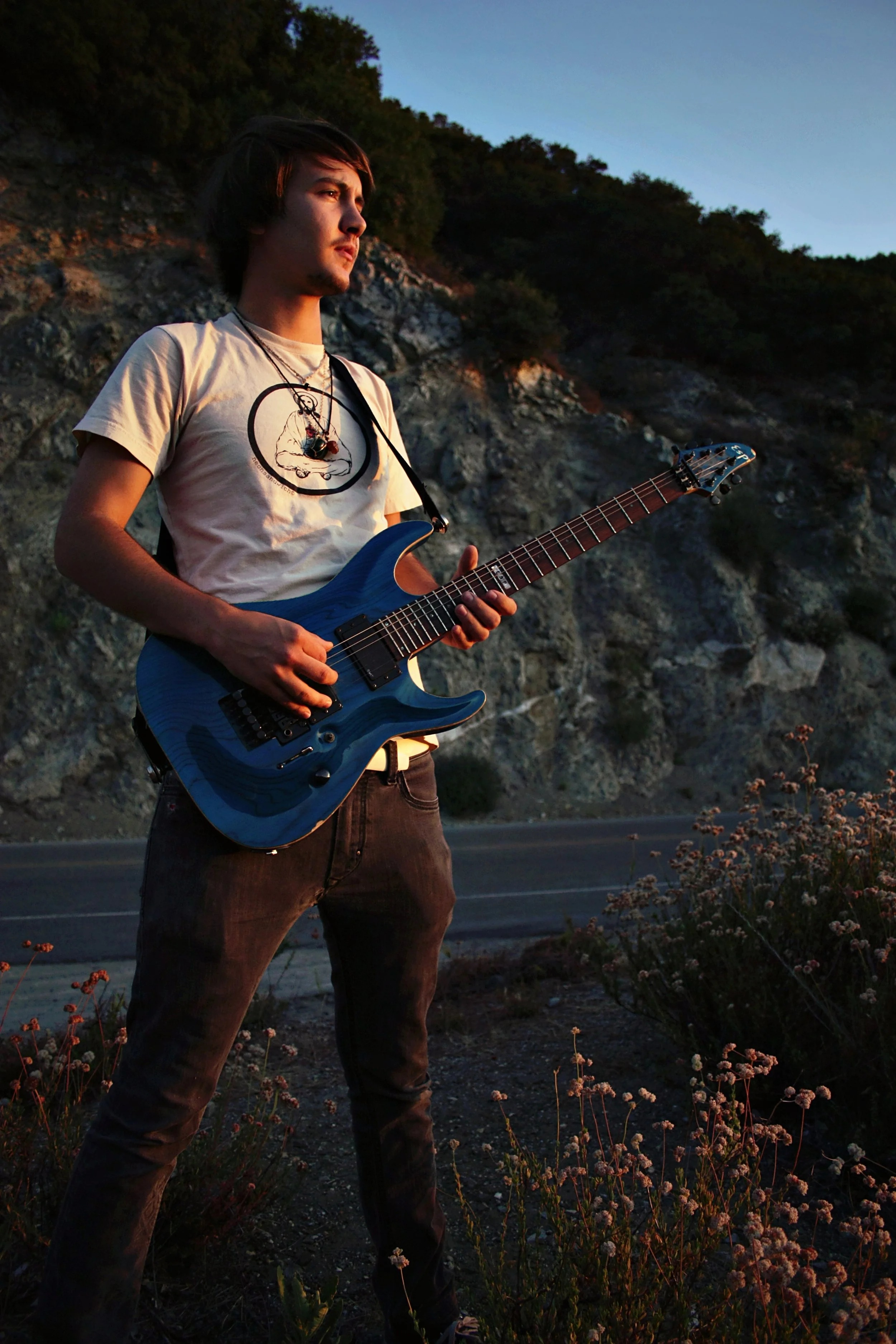 Young man with dark hair standing outdoors in the evening, holding a blue electric guitar, with a hillside and trees in the background.