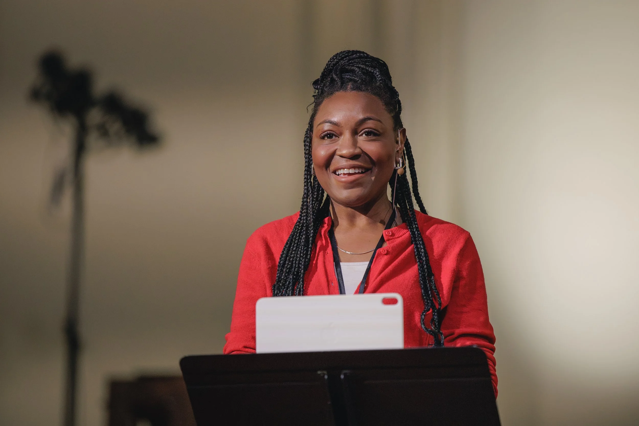 A woman with braided hair and earrings smiling while sitting at a table with a laptop, wearing a red top, with a blurred cross in the background.
