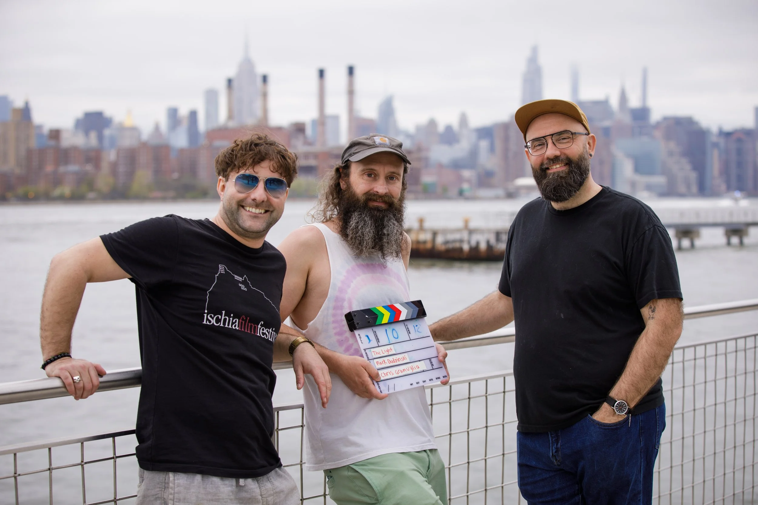 Three men smiling on a waterfront with the New York City skyline in the background, holding a film slate for a film production.