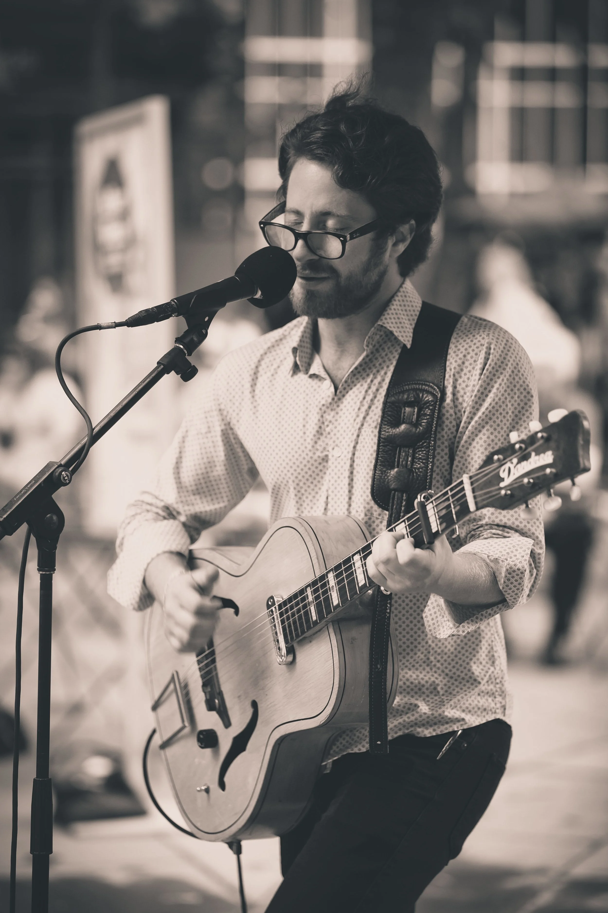 A man with glasses and a beard playing an acoustic guitar and singing into a microphone on a street.