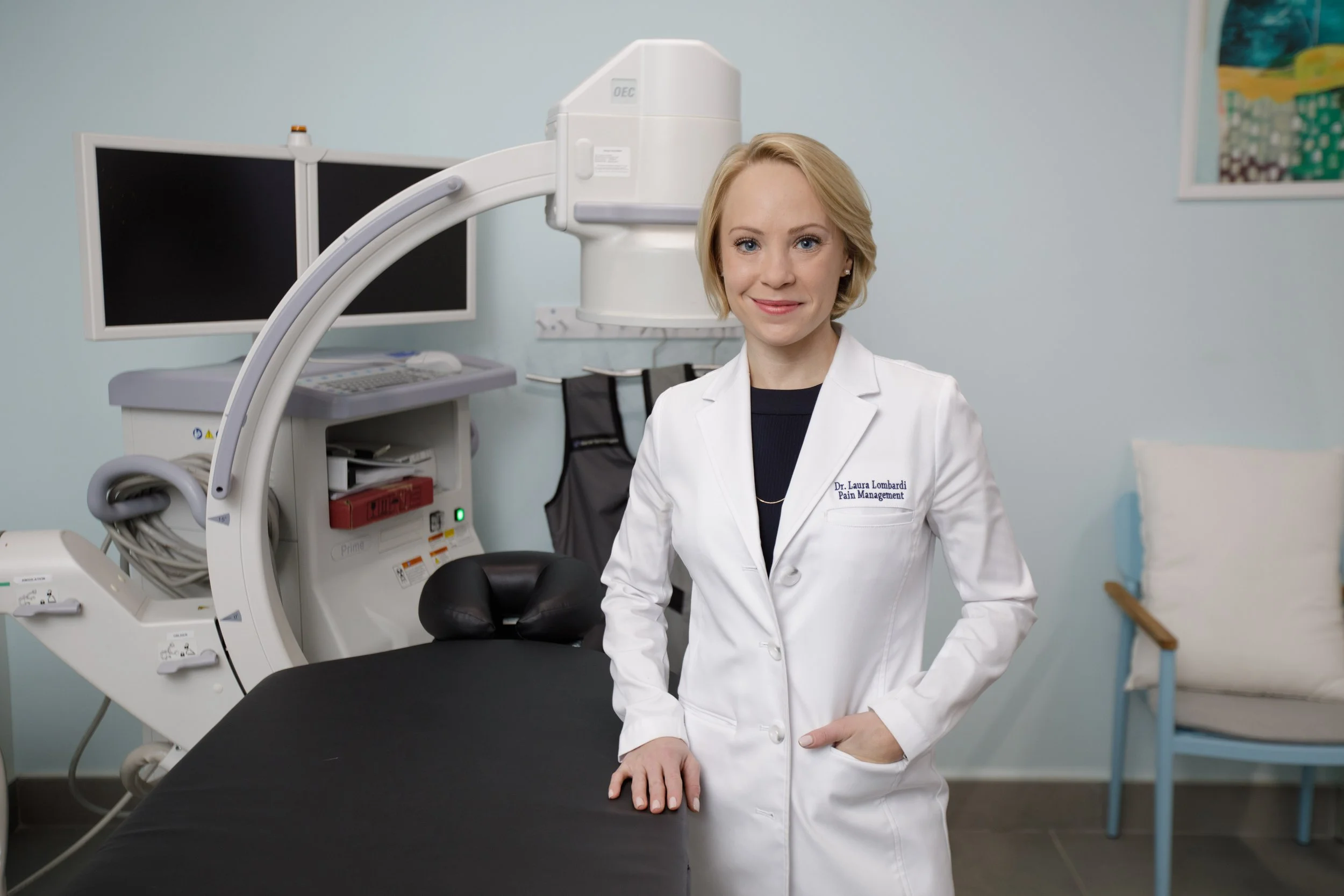 A woman in a white lab coat standing in a medical room with a CT scanner in the background.