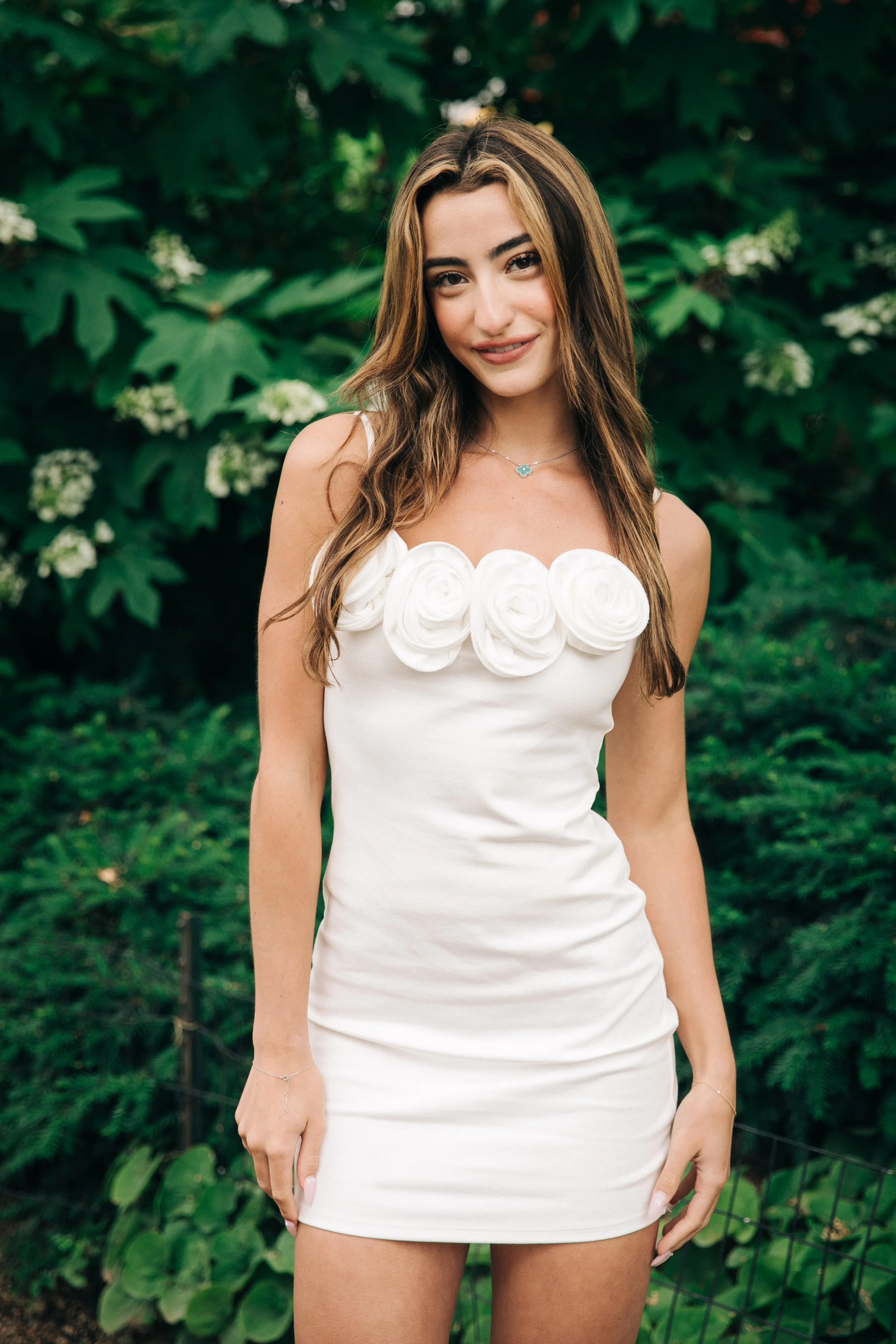 A young woman in a white dress with floral embellishments on the bodice, standing outdoors in front of green foliage.
