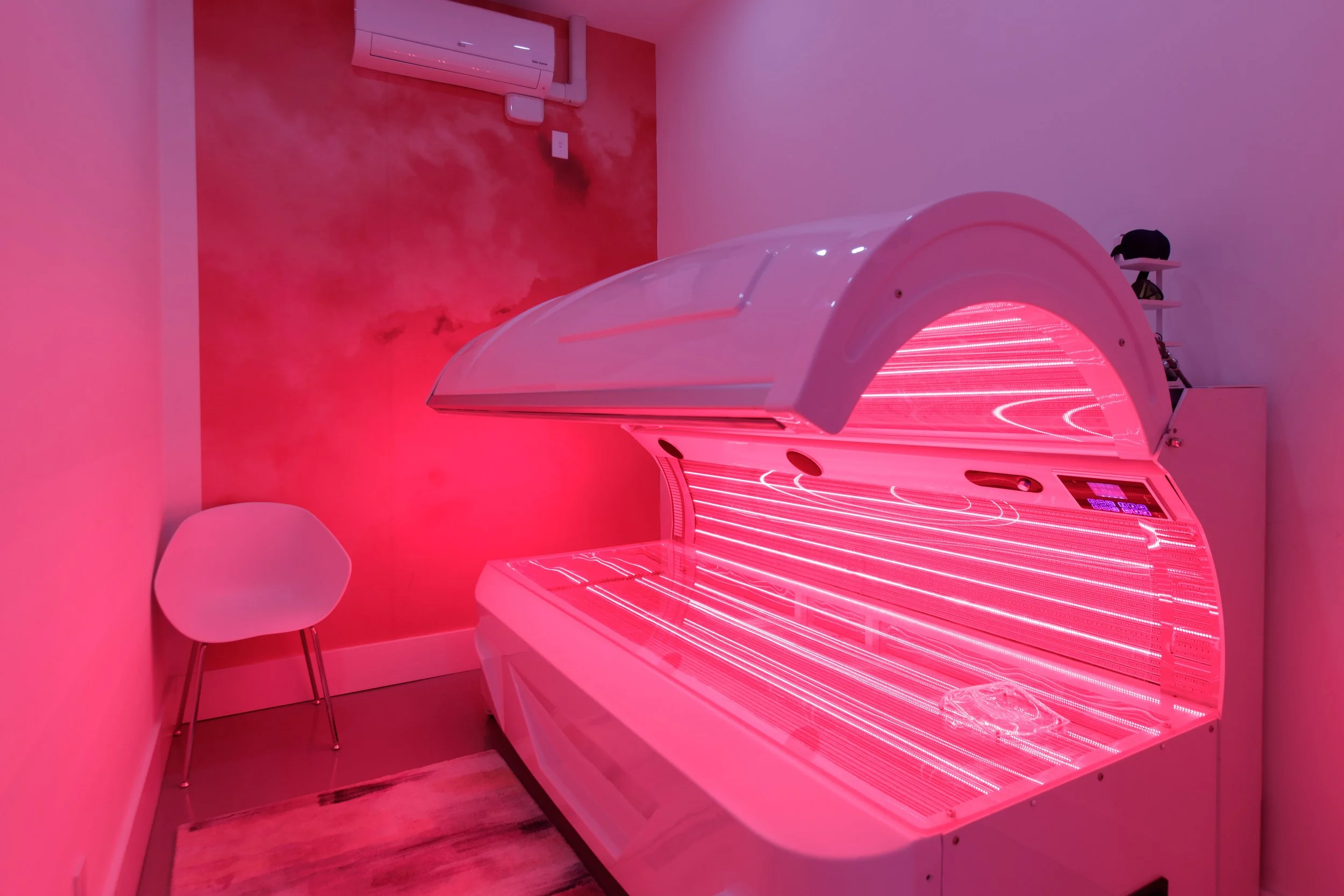 A pink indoor tanning bed emitting bright pink light in a room with pink walls, one white chair with metal legs, a shelf with a black hat, and an air conditioning unit on the wall.