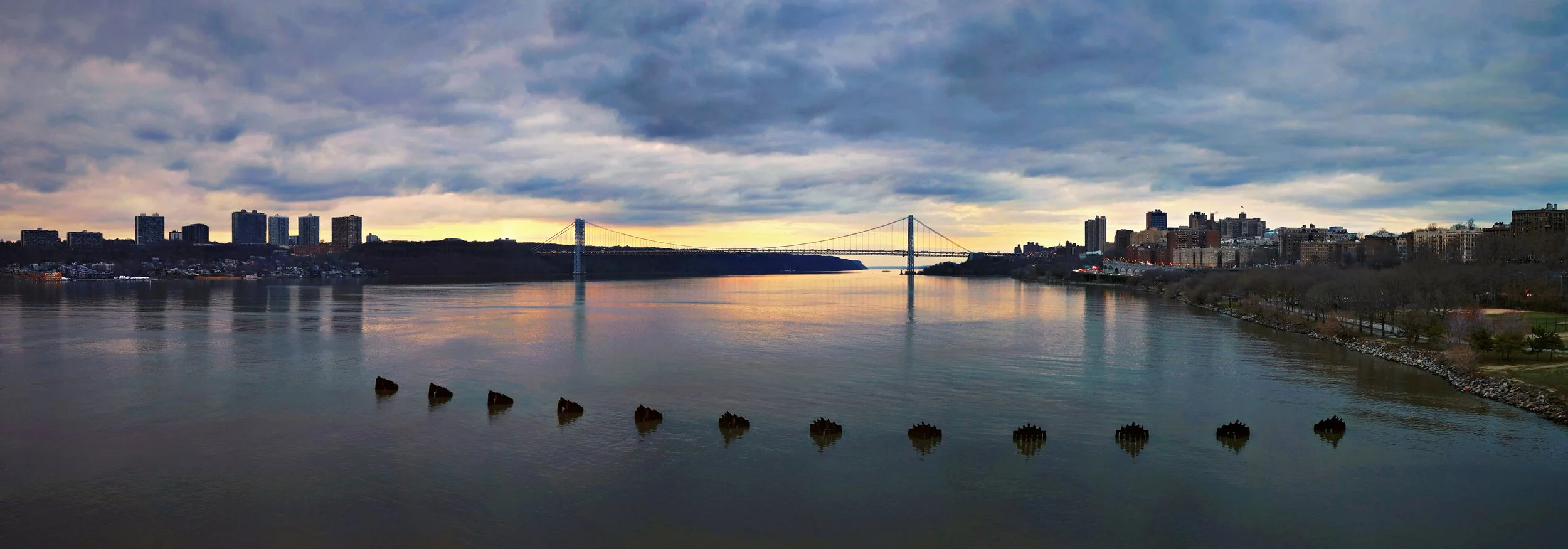 A wide river with a city skyline and a large suspension bridge in the background under a cloudy sky during sunset.
