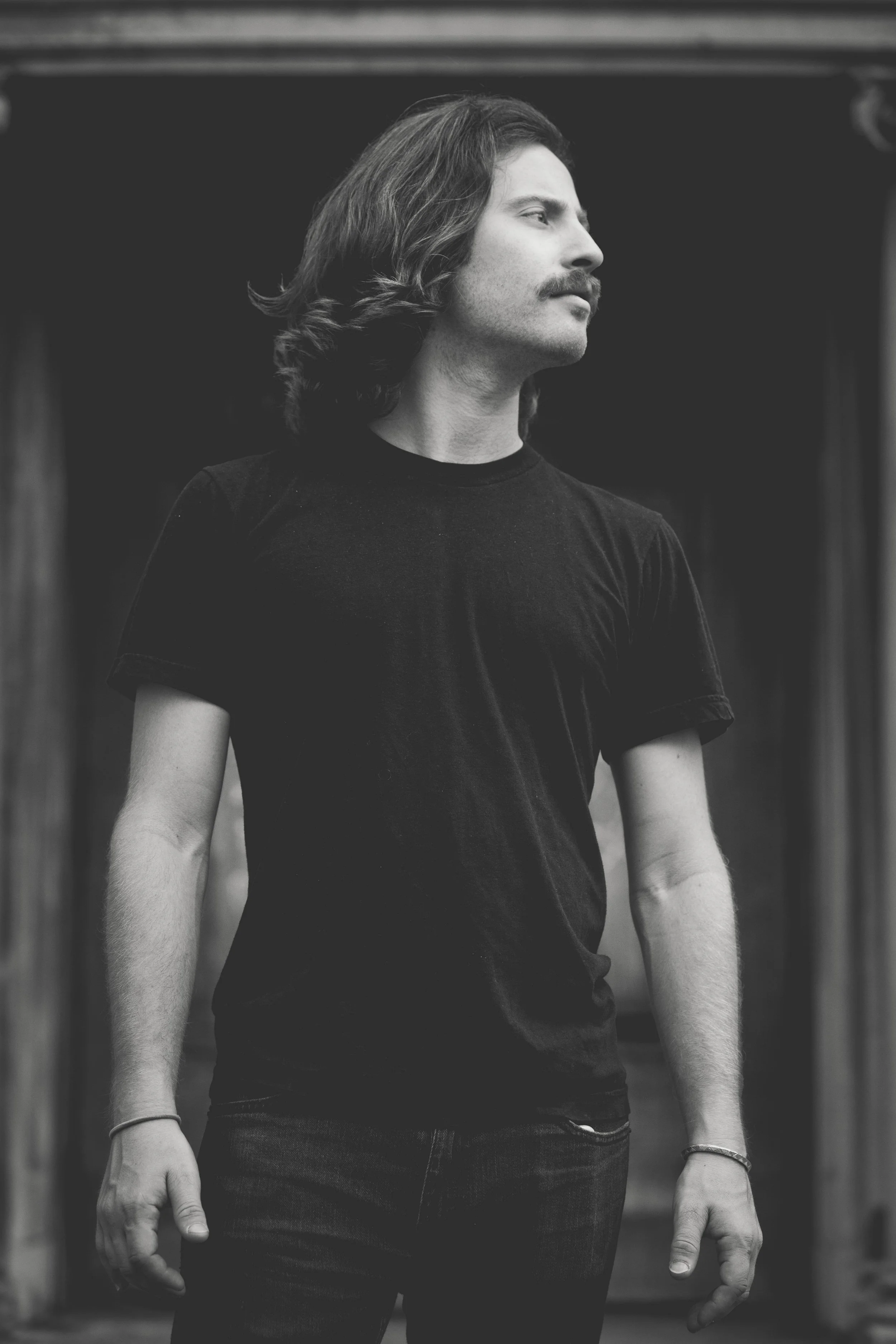 Black and white photo of a man with shoulder-length hair and a mustache, wearing a dark t-shirt, standing outdoors.