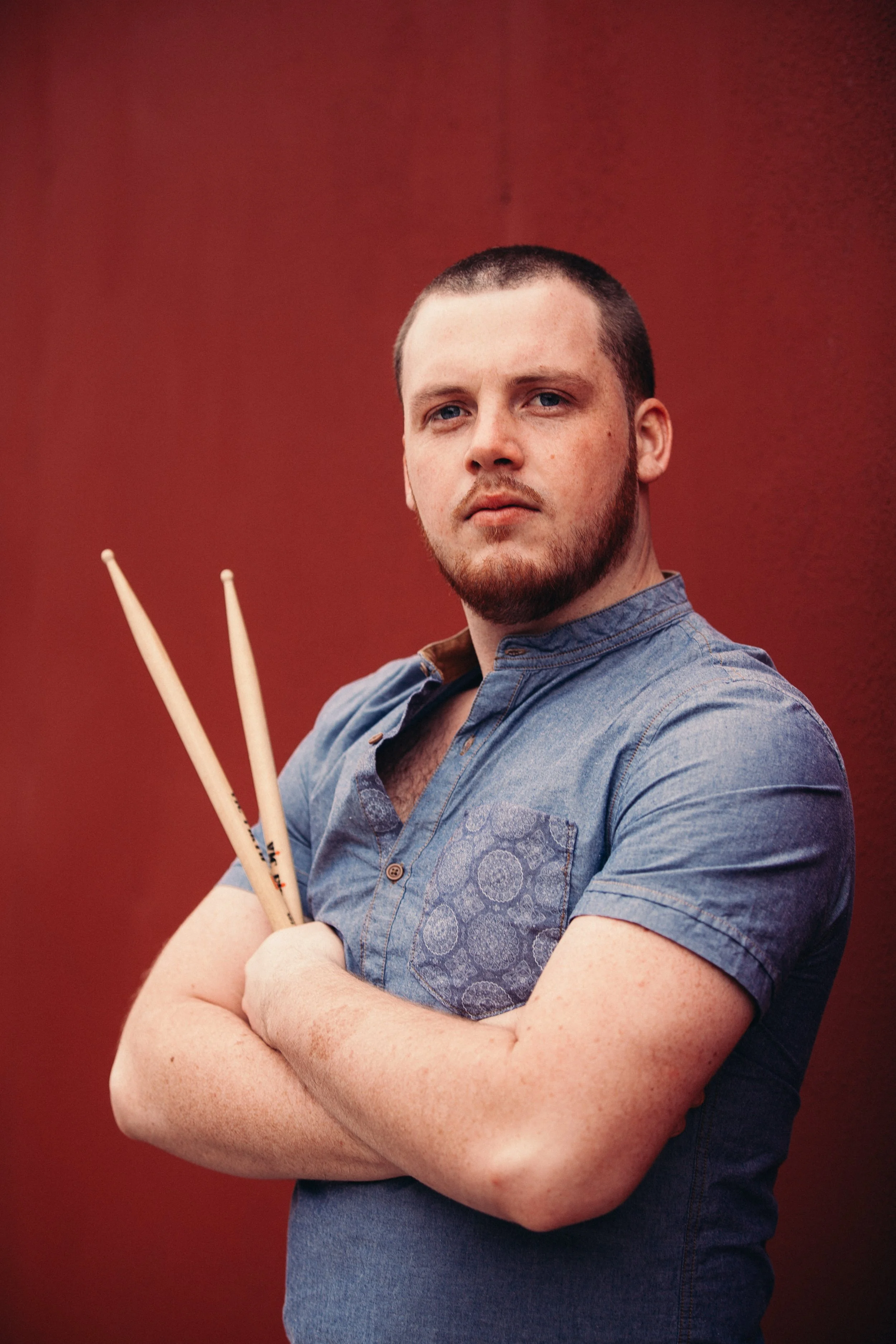 A man with short dark hair and a beard, wearing a blue shirt, posing with arms crossed in front of a red background, holding two drumsticks.