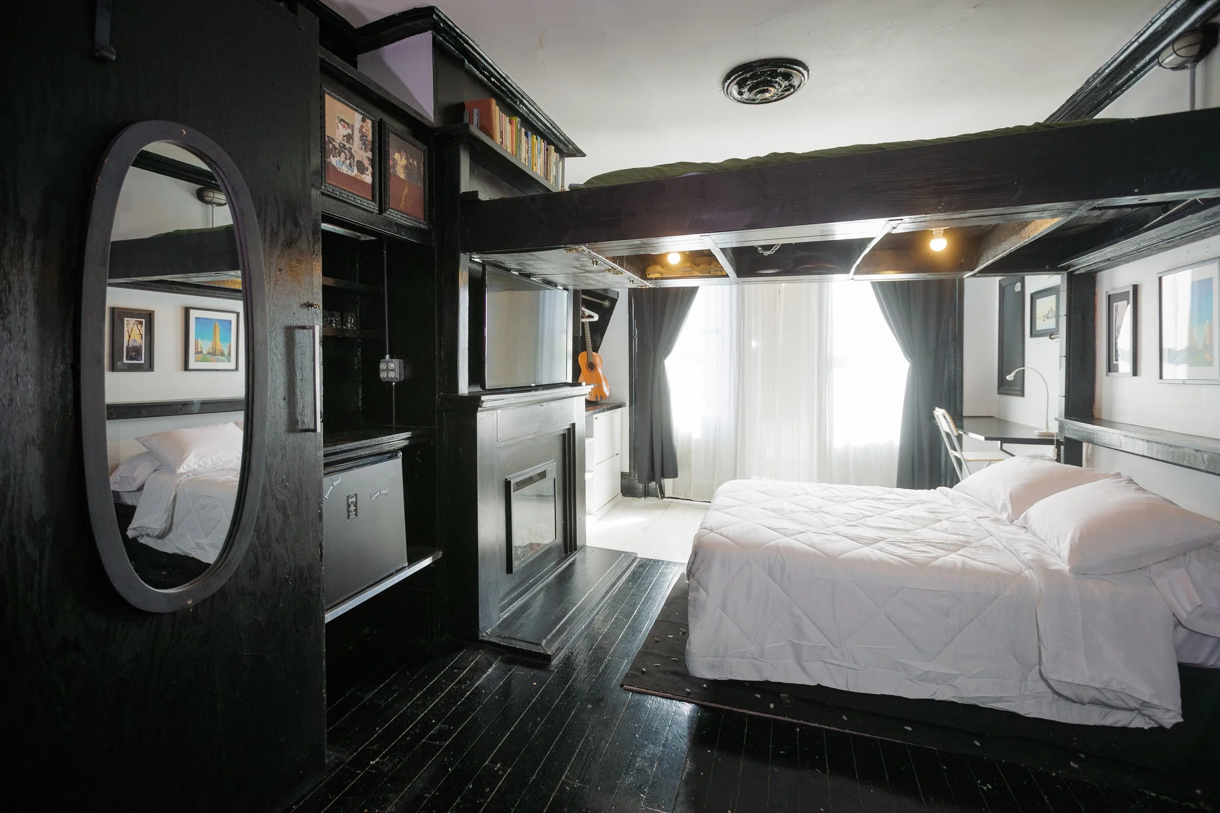 A bedroom with black walls and black wooden floors. There are two beds with white bedding, and a large window with white curtains. A guitar is on a stand near the window. The image is reflected in a tall mirror on a black wooden partition, which also