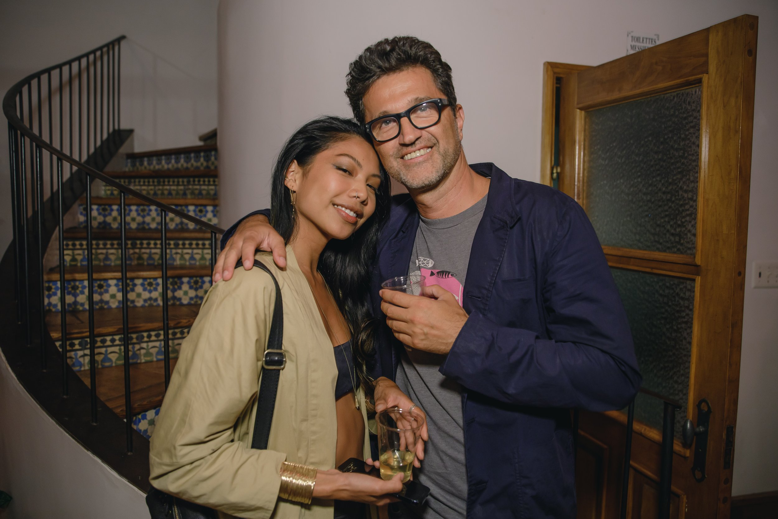 A woman and a man are smiling and hugging at a social gathering, holding drinks, standing near a staircase with colorful tile carpet and a wooden framed glass door.