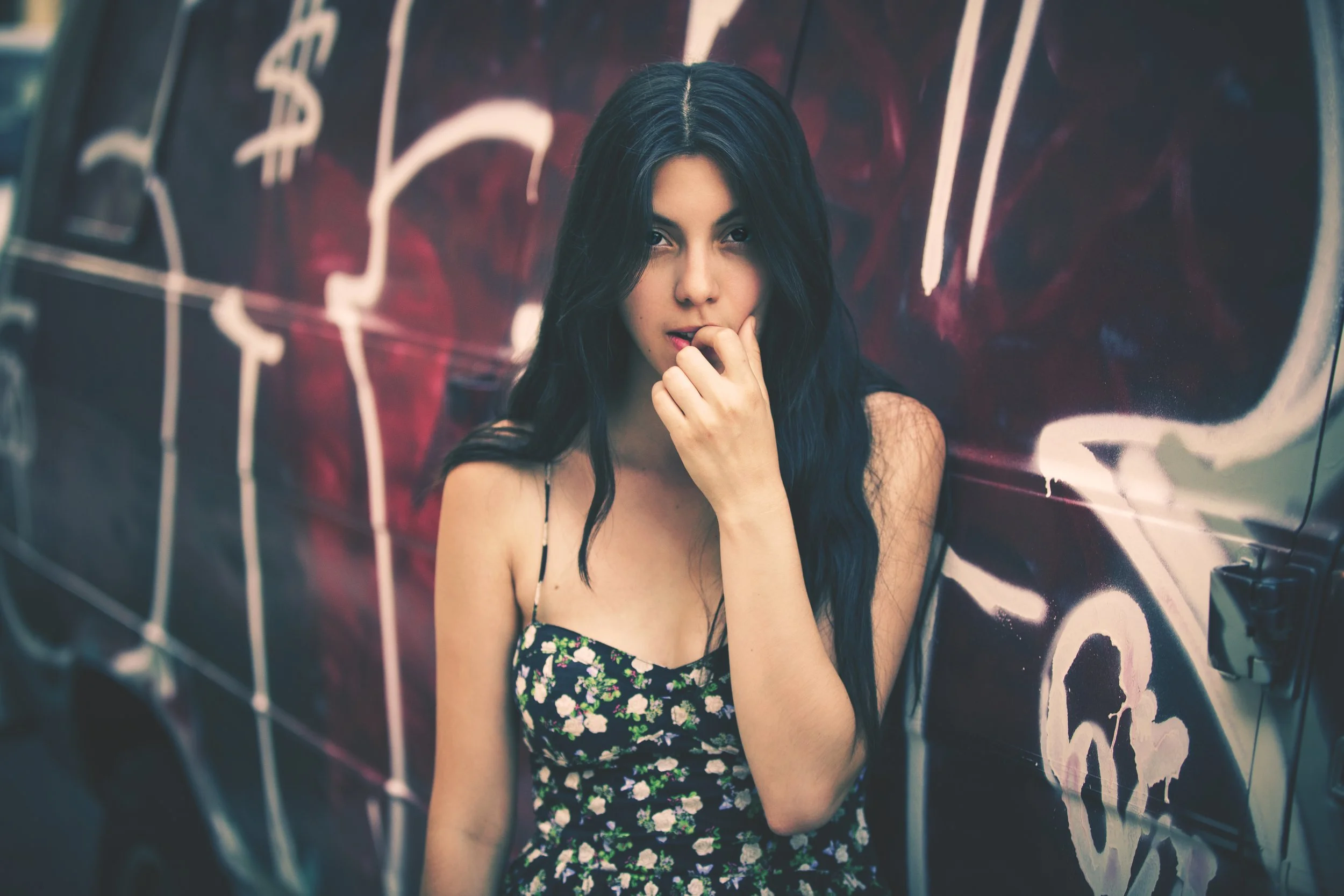 A young woman with long dark hair and a floral dress posing against a graffiti wall.