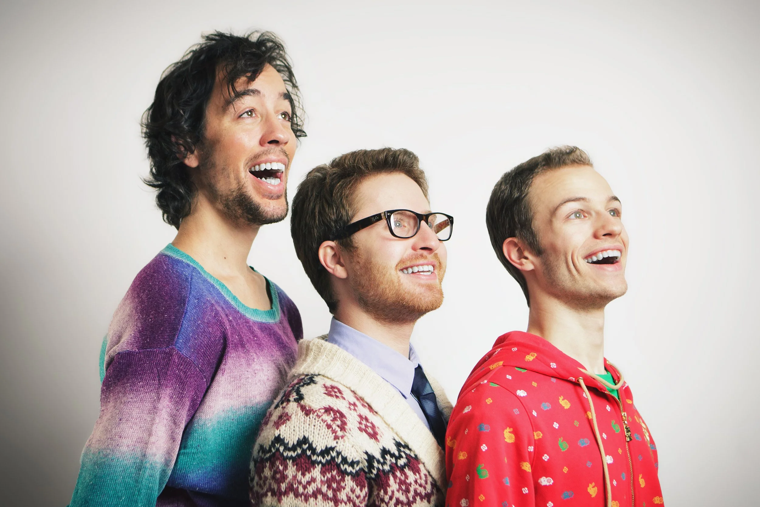 Four young men standing side by side, looking upward with happy expressions, wearing colorful sweaters and clothing, against a plain white background.