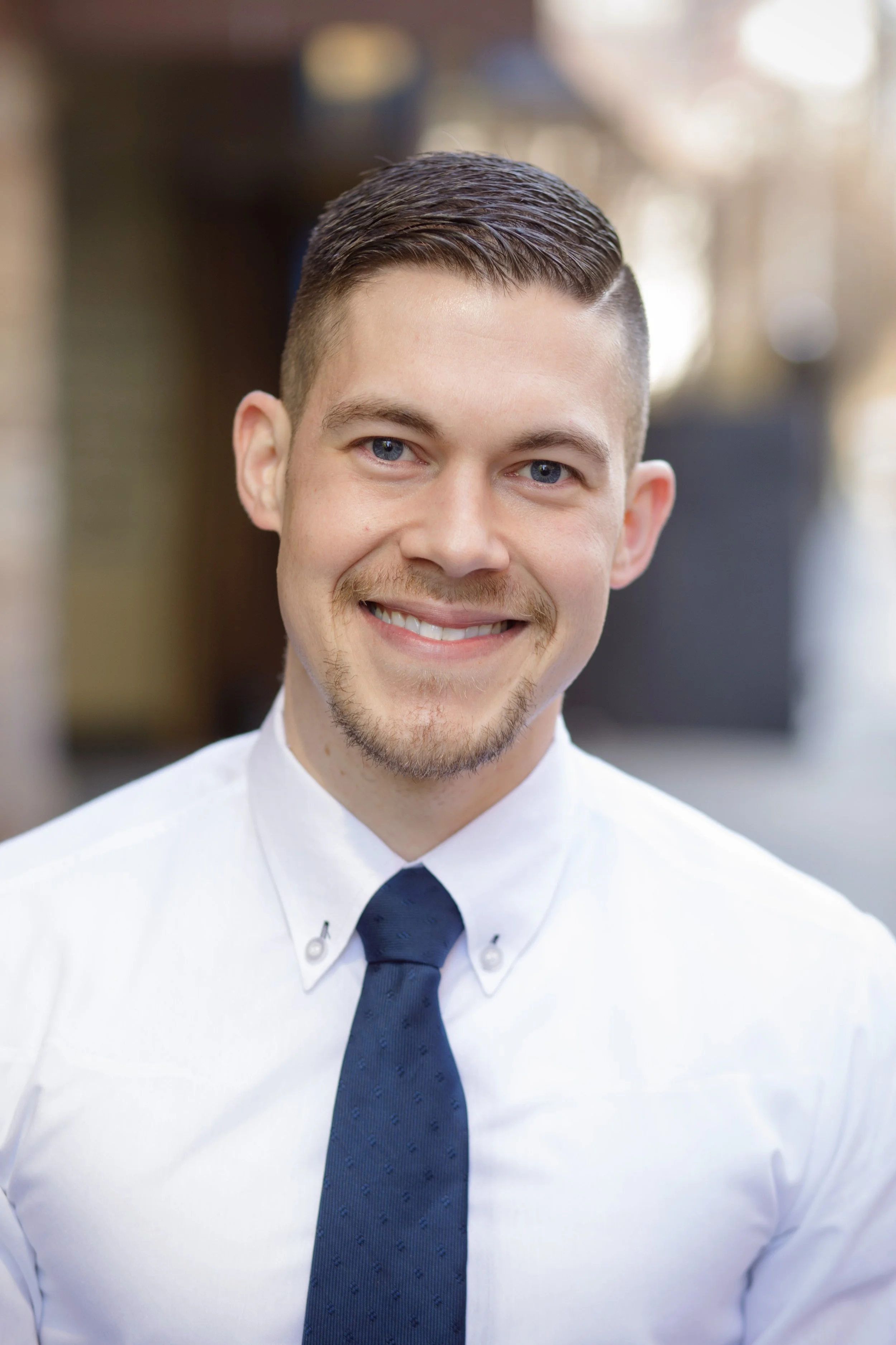 Close-up portrait of a young man with short dark hair, blue eyes, and a light beard, smiling, wearing a white shirt and a dark tie, with a blurred indoor background.