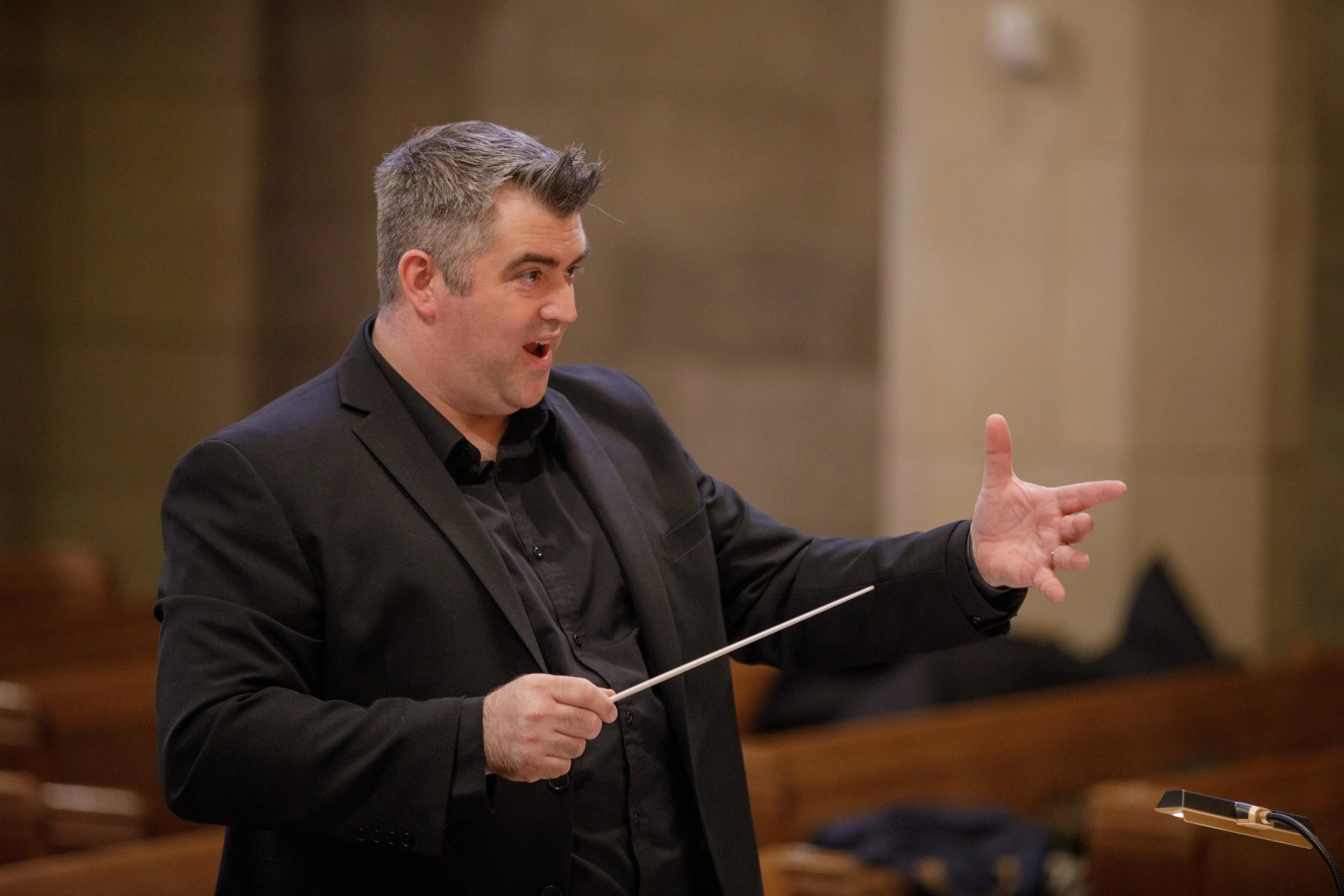 A man in a black suit and shirt conducting or presenting, holding a baton, in a formal setting.