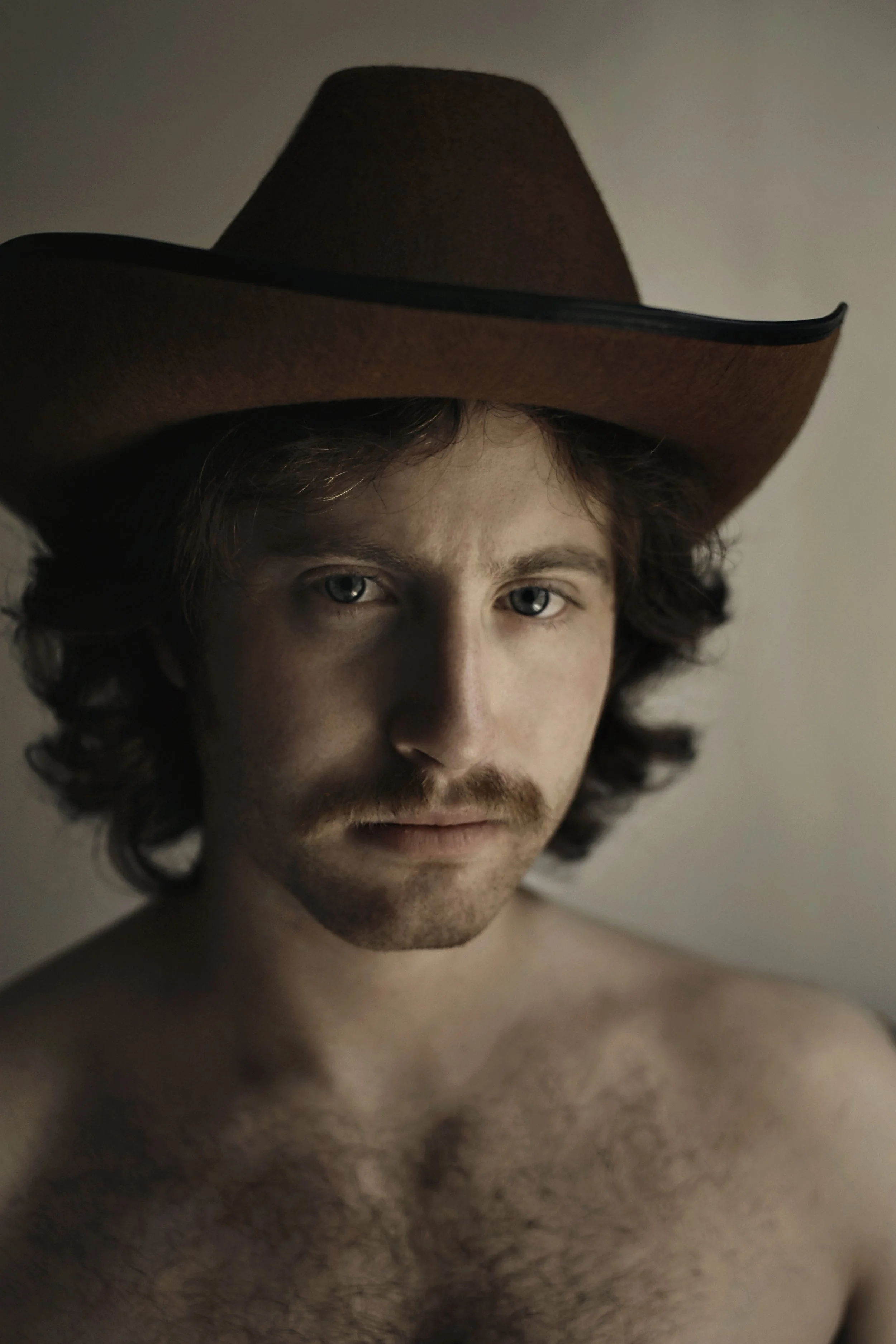 A young man with wavy hair and a mustache wearing a large brown cowboy hat with a black band.