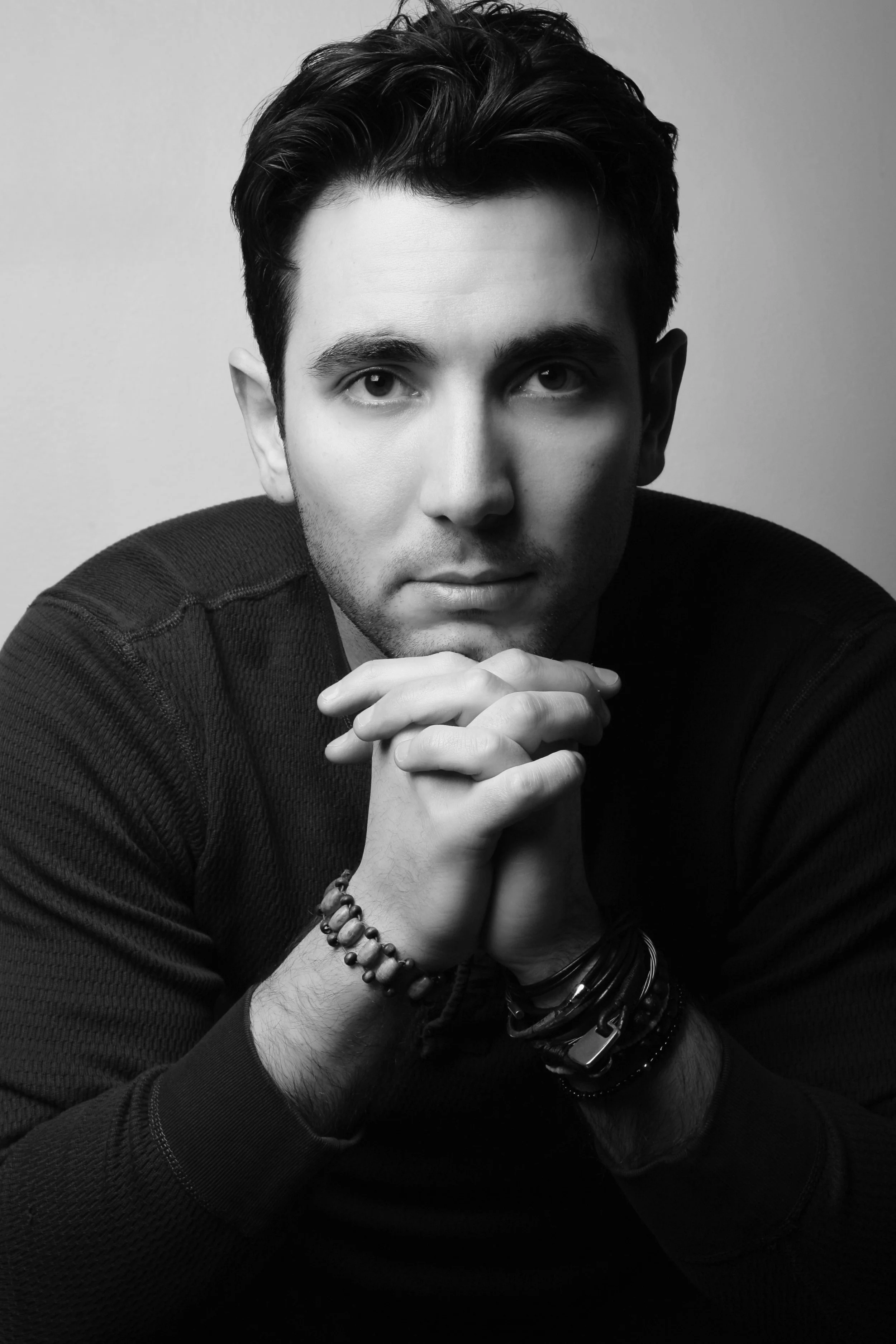 Black and white portrait of a young man with dark hair, wearing a long-sleeve shirt, with hands clasped together under his chin, wearing bracelets, and looking directly at the camera.