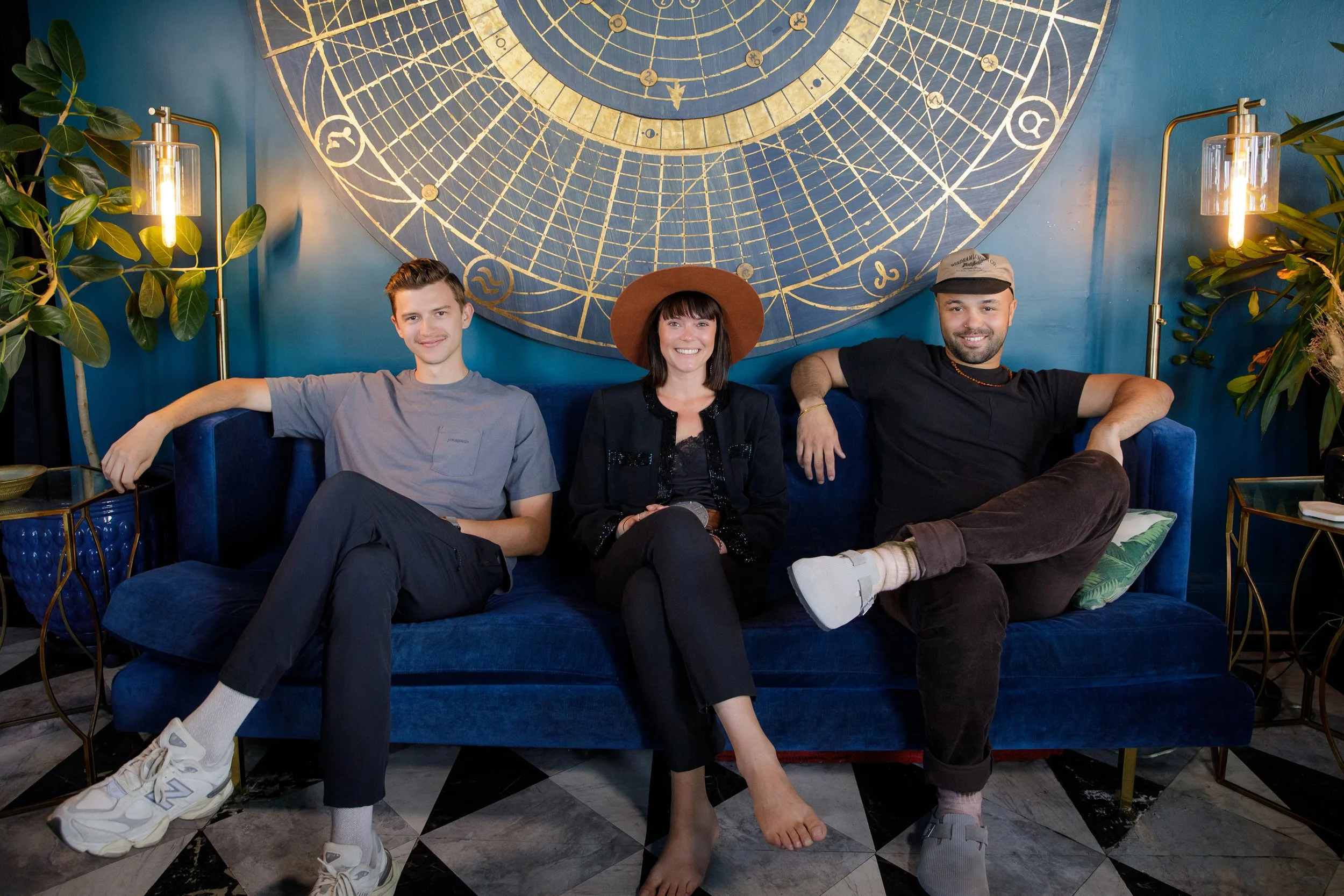 Three young adults sitting on a blue velvet sofa in a decorative room with a zodiac chart on the wall behind them. The person in the middle is wearing a wide-brimmed hat, the person on the right is wearing a cap, and the person on the left is dressed