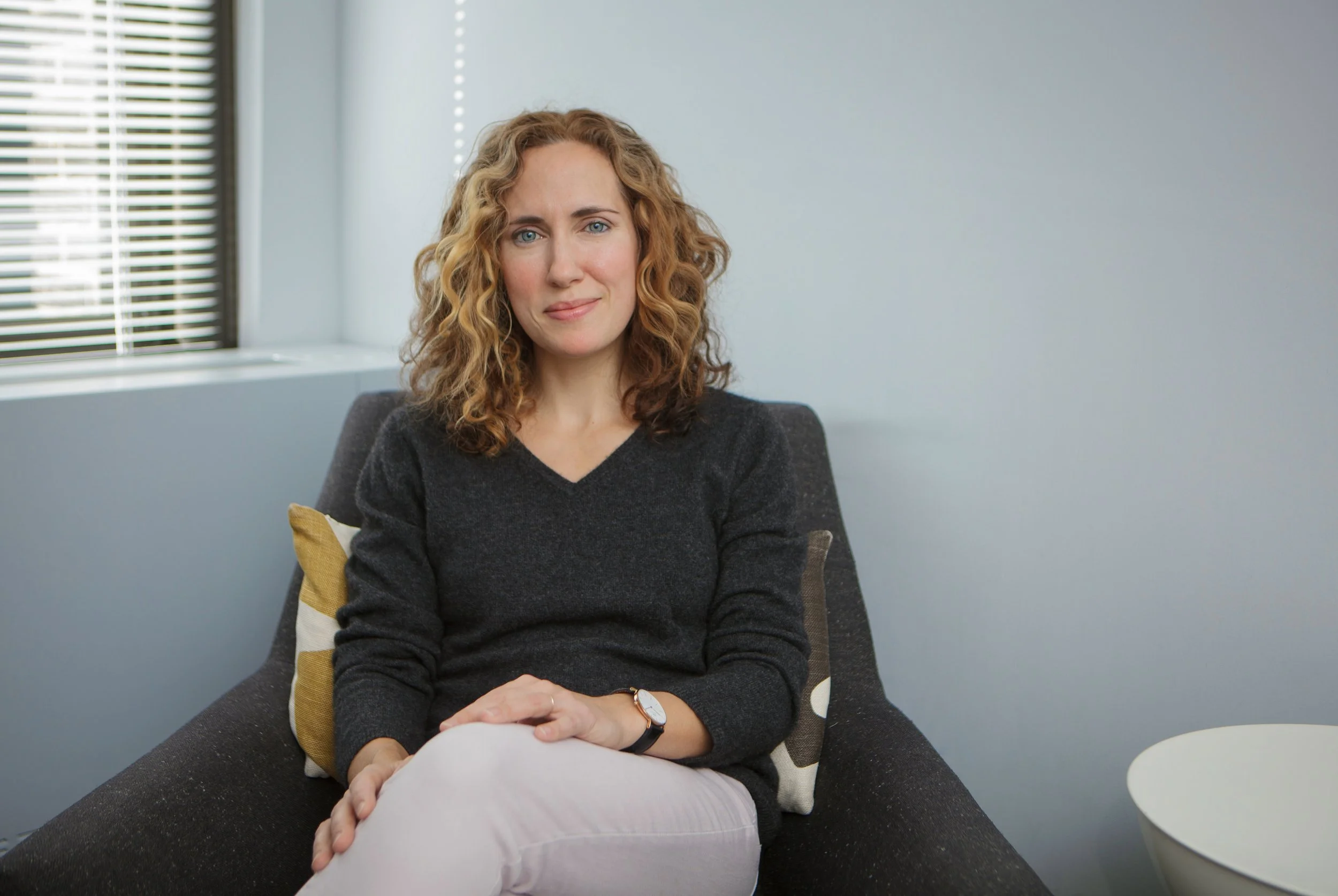 A woman with curly blonde hair and blue eyes sitting on a dark gray sofa in a modern office, smiling softly at the camera.