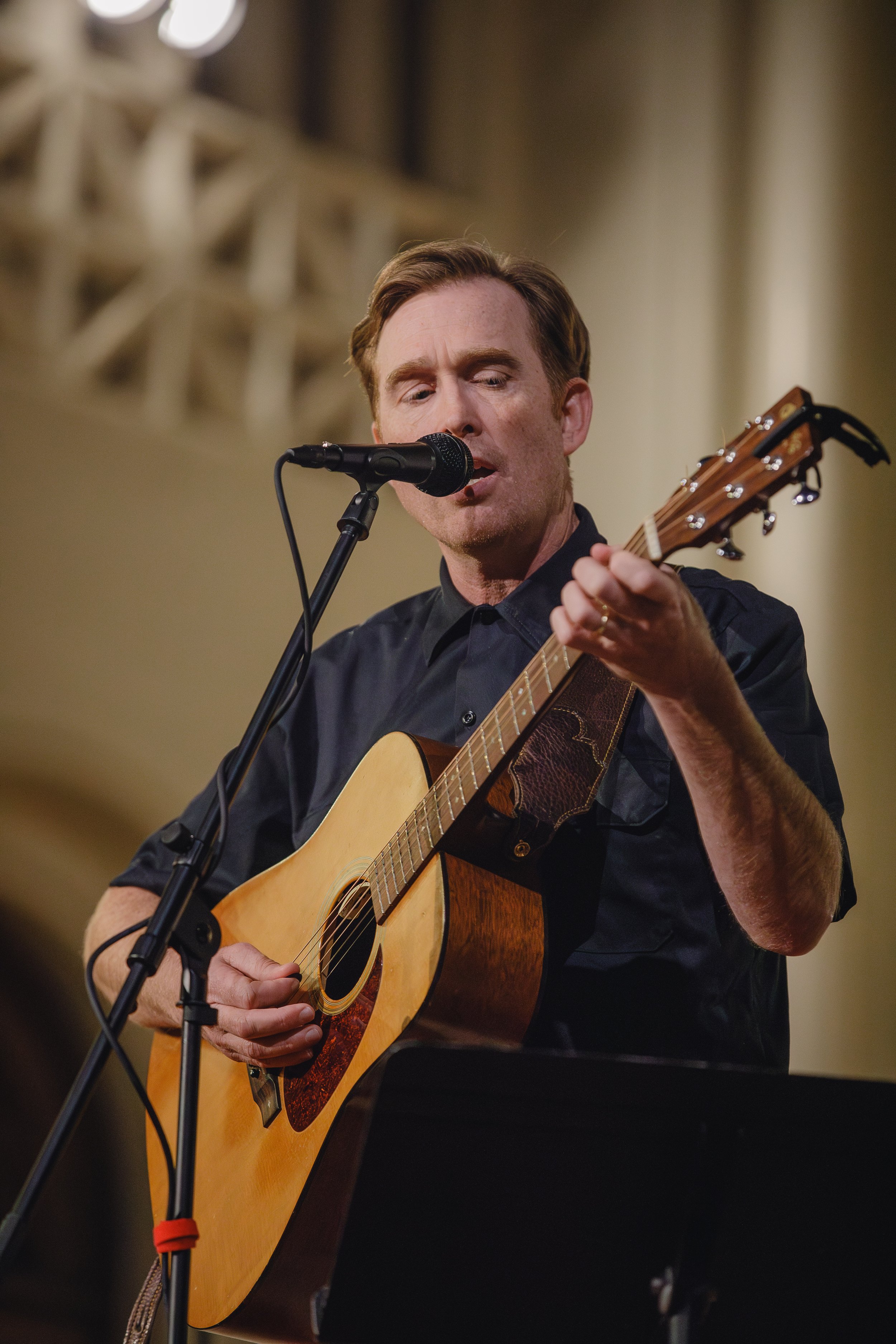 A man with reddish hair and wearing a black shirt plays acoustic guitar and sings into a microphone during a performance.