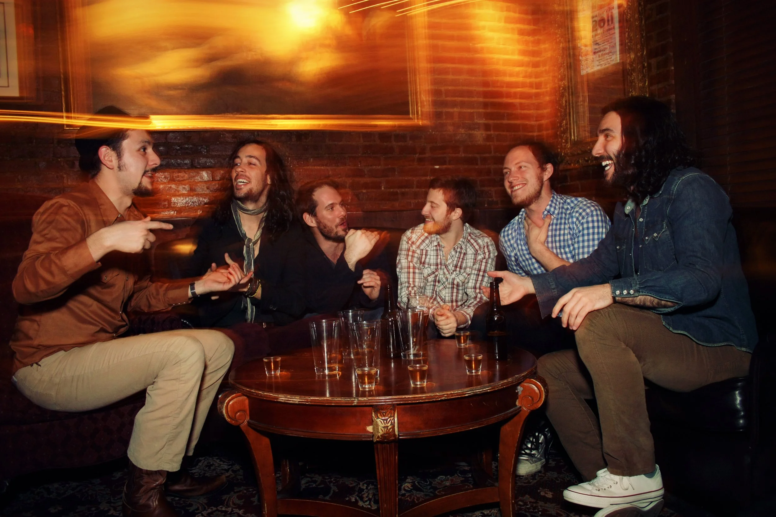 Group of six men sitting around a wooden table in a bar or pub, engaged in conversation, some holding shot glasses. The background features a brick wall and framed artwork, with warm lighting creating a lively atmosphere.