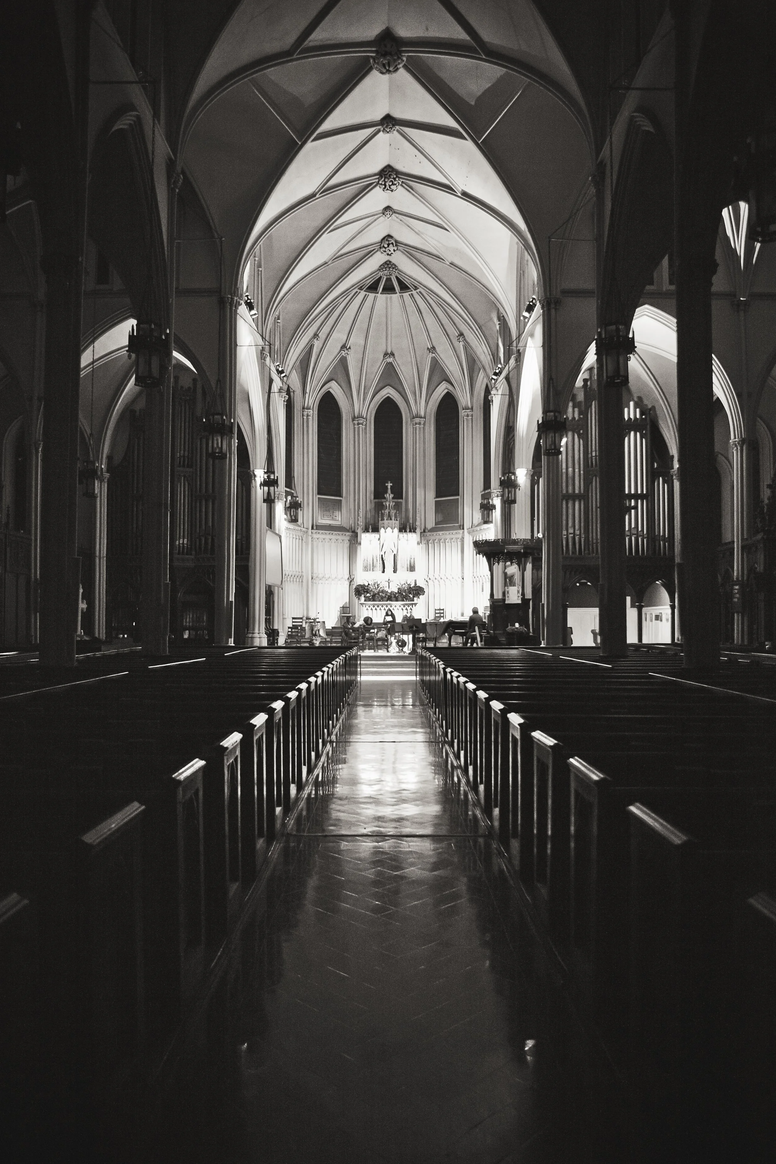 Interior of a Gothic-style church with pointed arches, high vaulted ceilings, and wooden pews arranged in rows leading to the altar at the front. The church is illuminated from the back near the altar, with some silhouetted figures visible.
